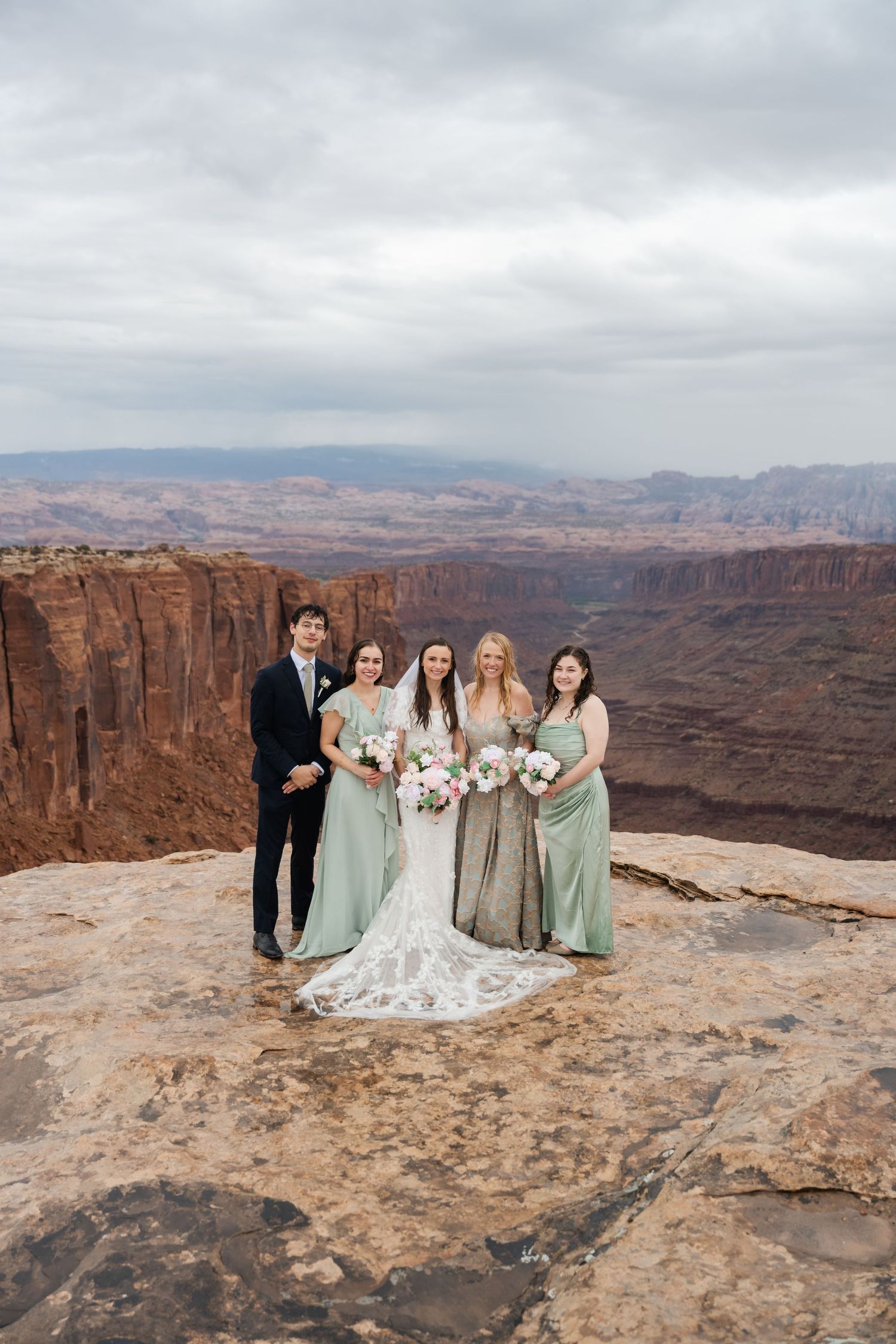 Wedding party in sage green dresses gathered at scenic desert canyon viewpoint.