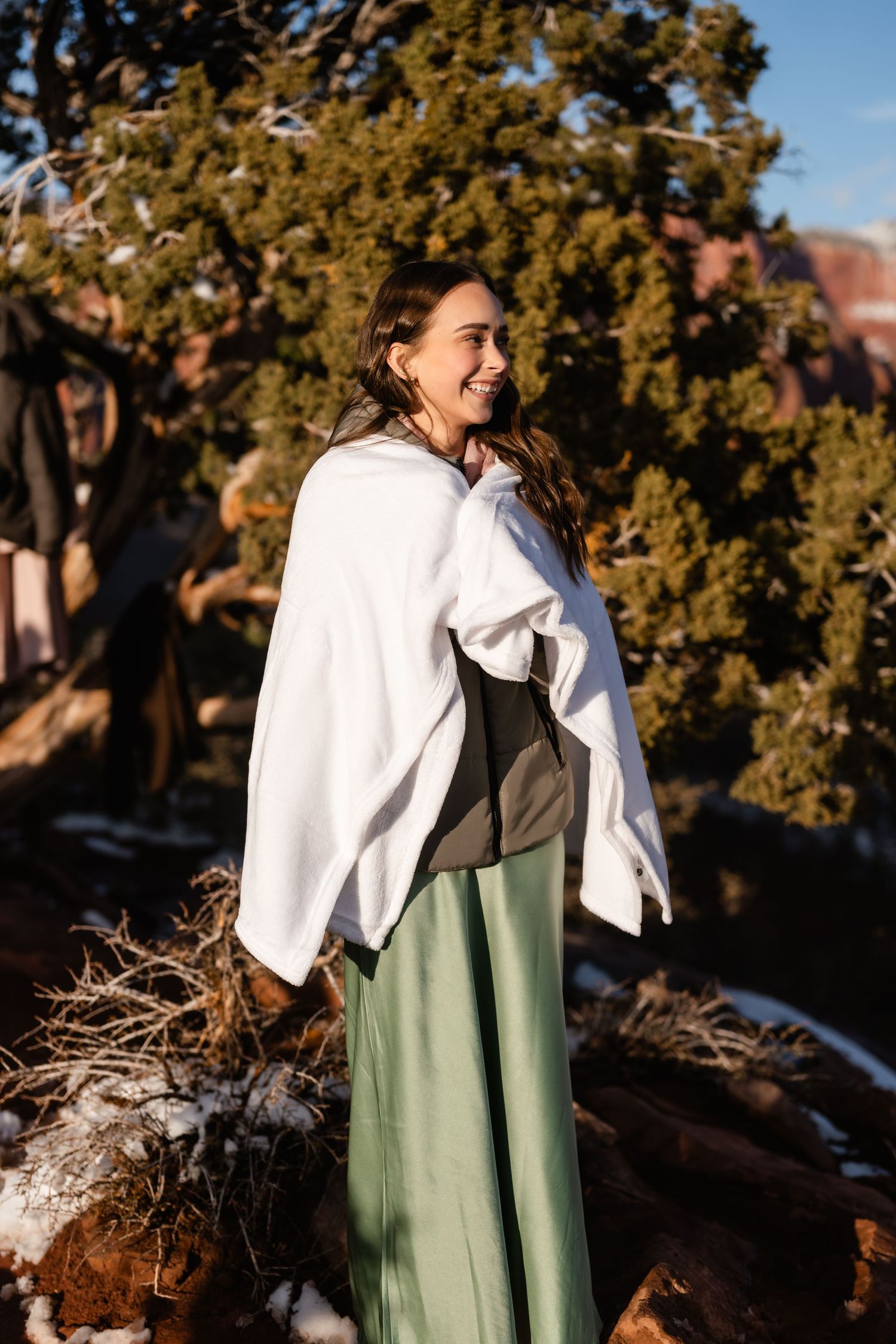 Person wearing a white shawl and sage green dress stands against a desert mountain backdrop.