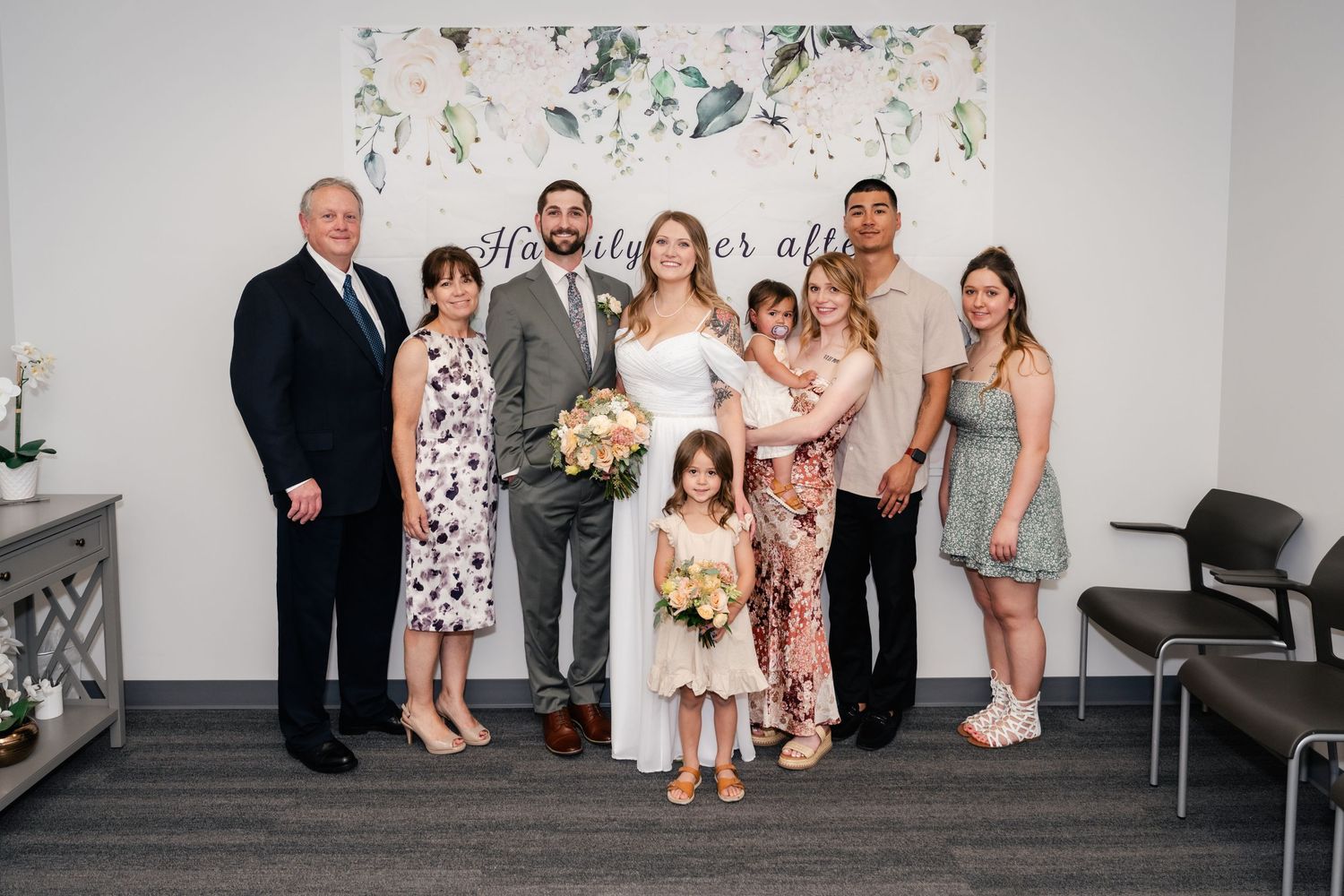 Wedding party poses together in front of white floral wall backdrop at elegant indoor venue.