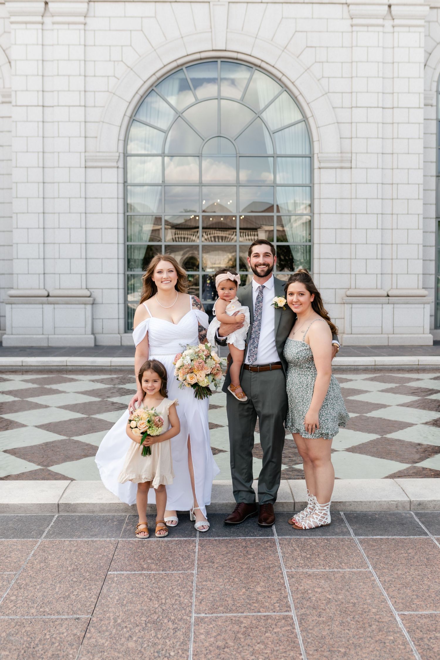 Wedding group stands in front of elegant white stone building with arched windows and checkered tile floor.