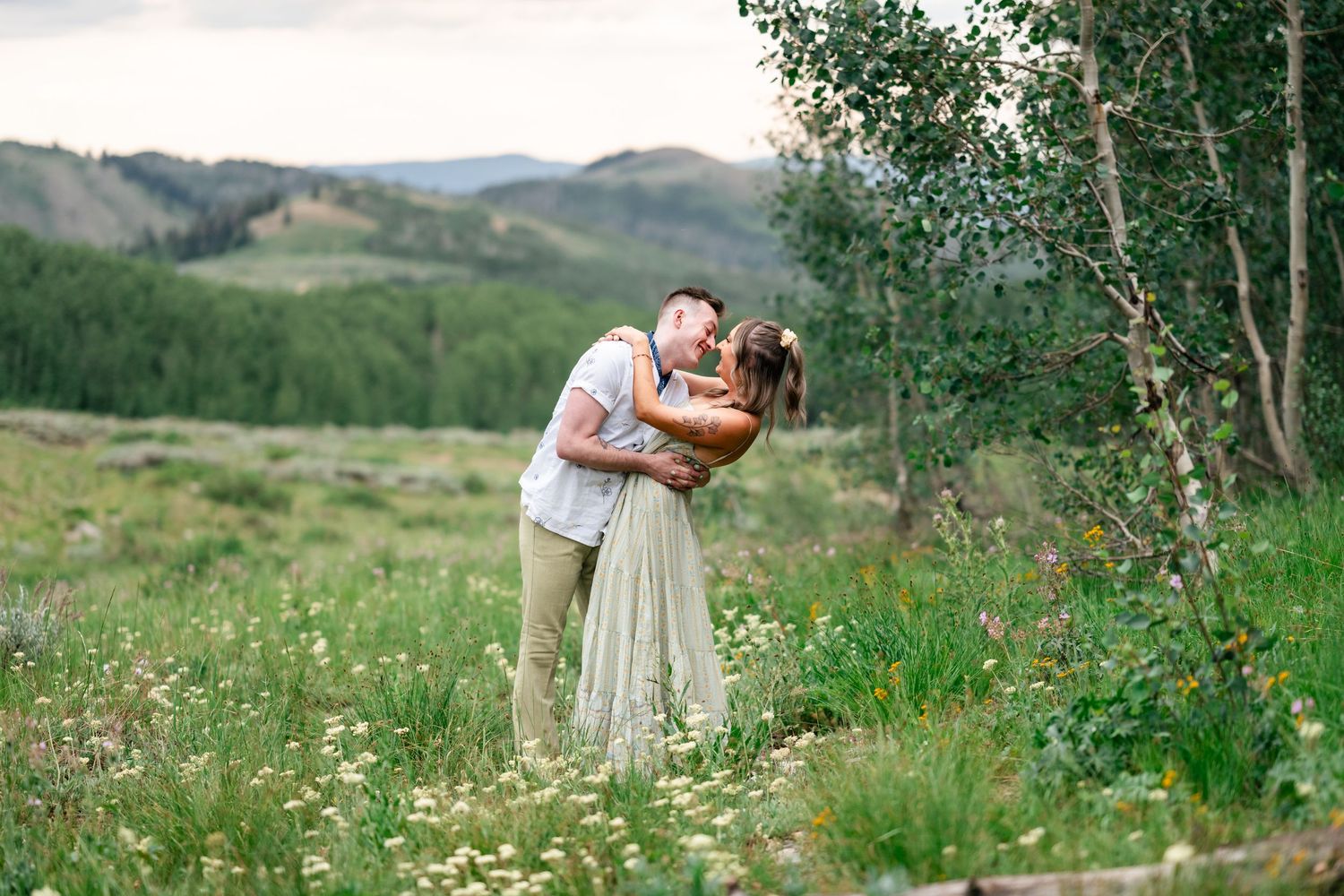 A romantic couple embraces in a mountain meadow surrounded by aspen trees and wildflowers on a summer day.