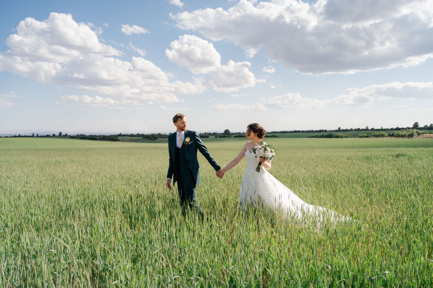 A couple in wedding attire walks hand in hand through a lush green wheat field under a bright cloudy sky.
