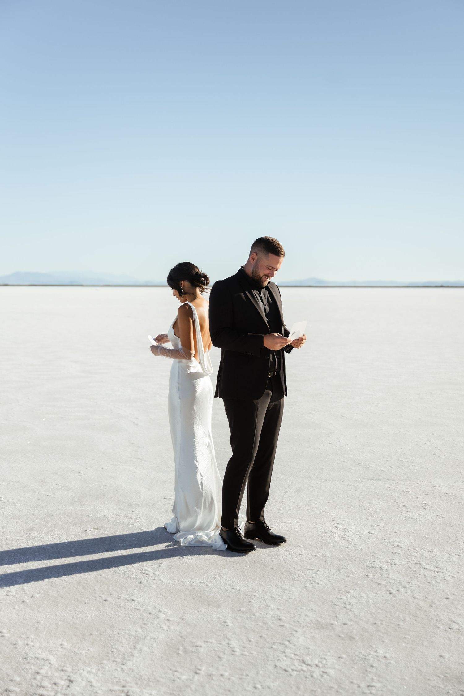 Couple in formal attire standing on salt flats during sunset.
