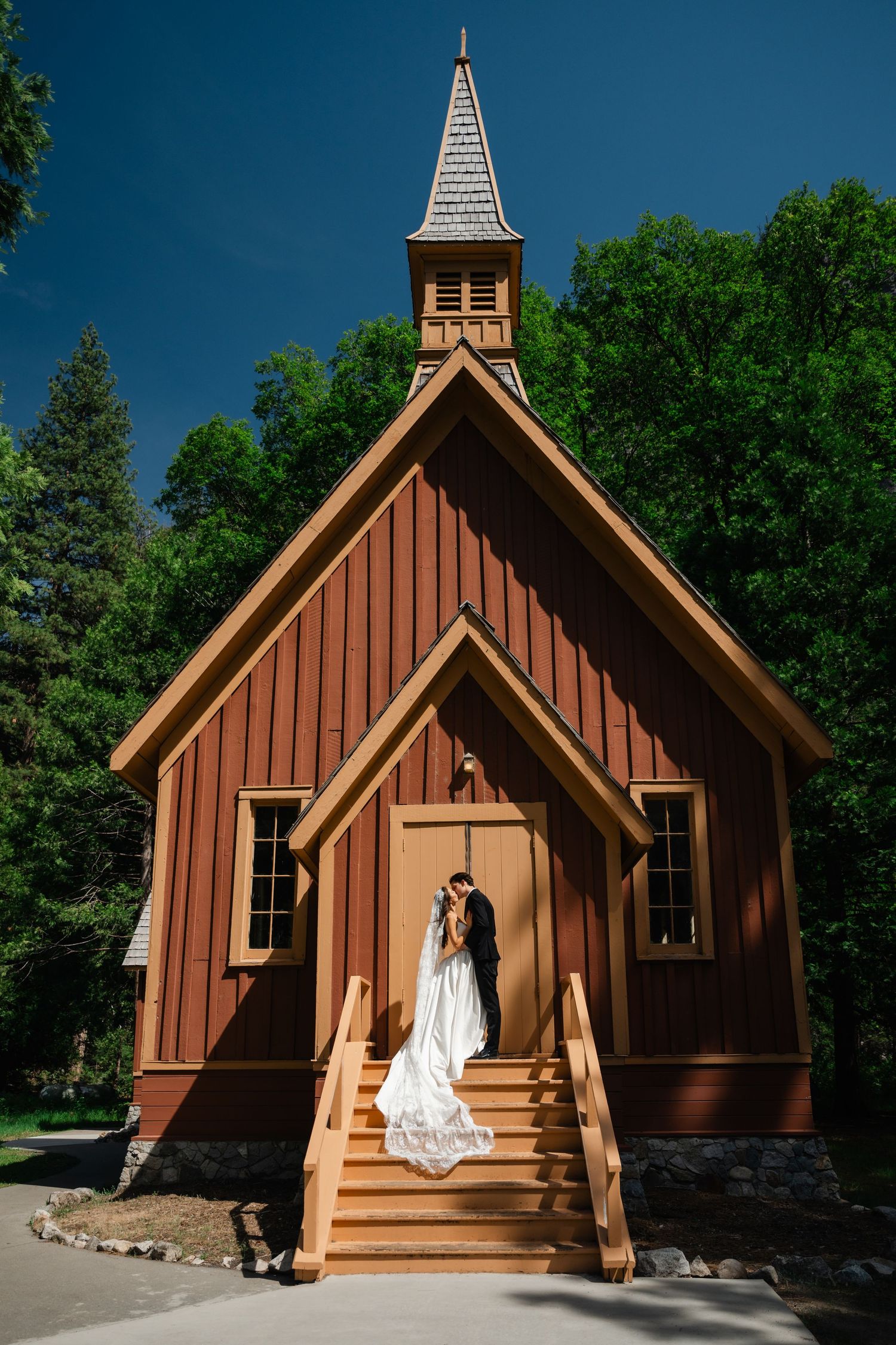 Bride and groom kissing on steps of rustic red wooden chapel with steeple surrounded by green trees under blue sky.