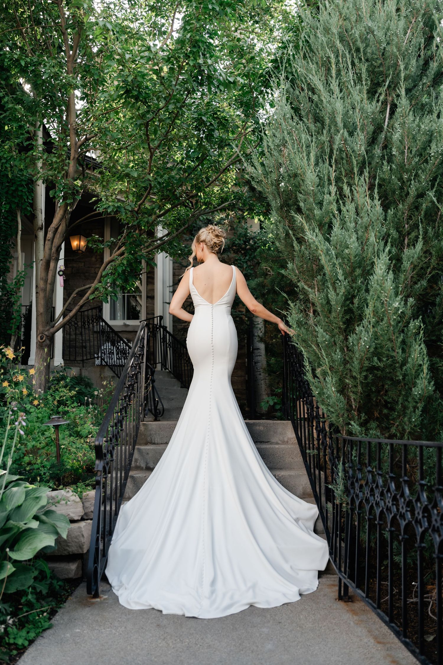 Bride in elegant white mermaid wedding gown with train poses on stone steps surrounded by lush greenery during their Park City micro wedding