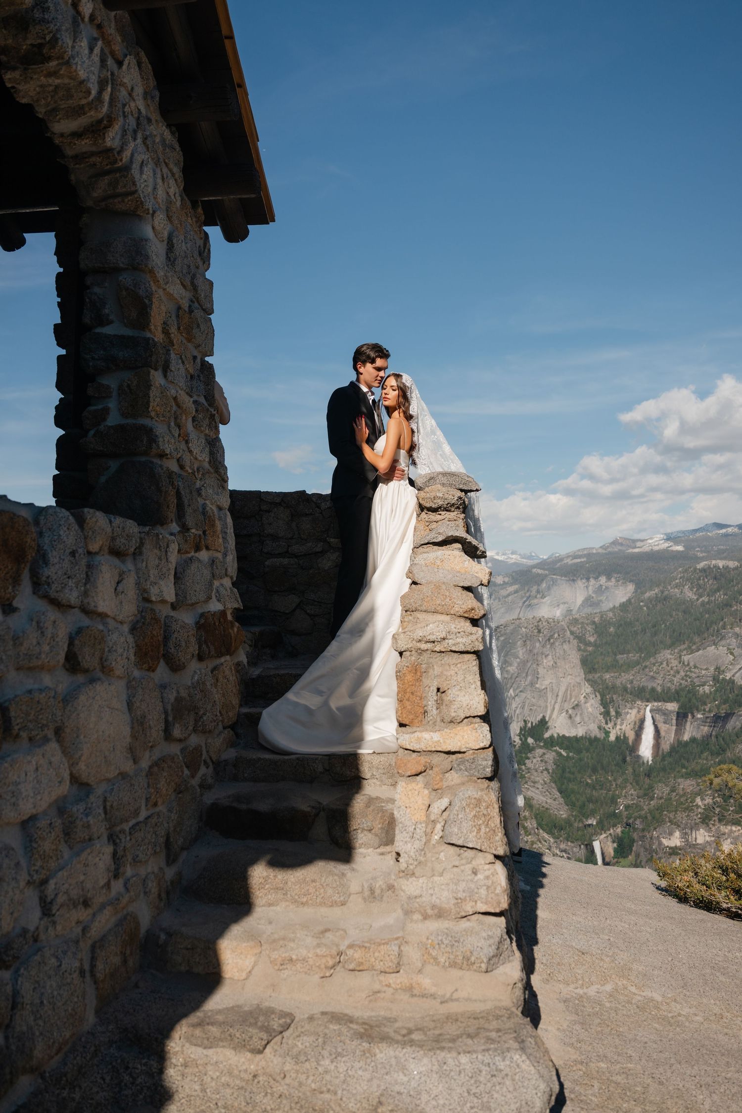 Bride and groom embrace on a stone overlook at Yosemite with waterfall and mountain views behind them.