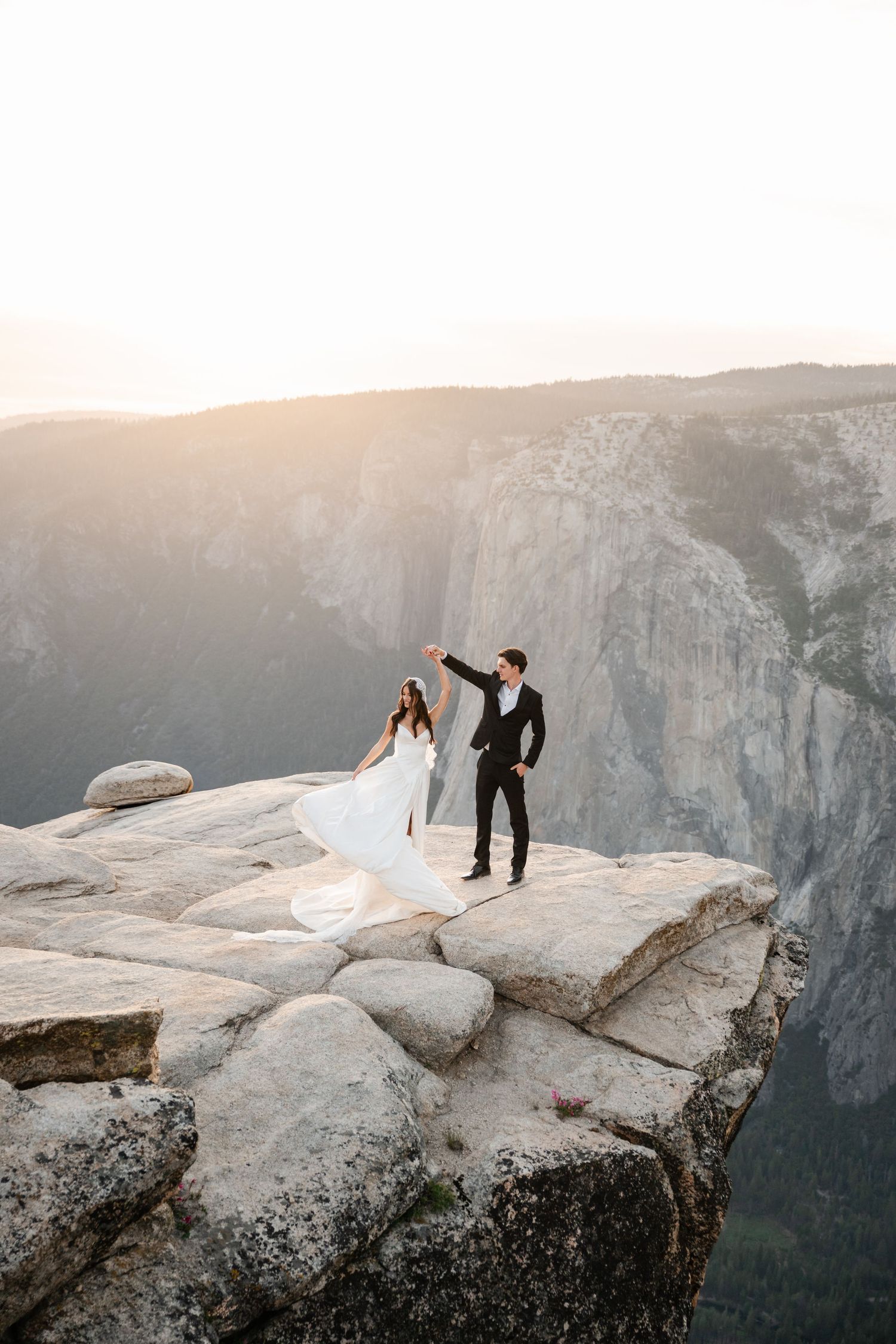 Bride in flowing white gown and groom dance joyfully on rocky mountain cliff at sunset.