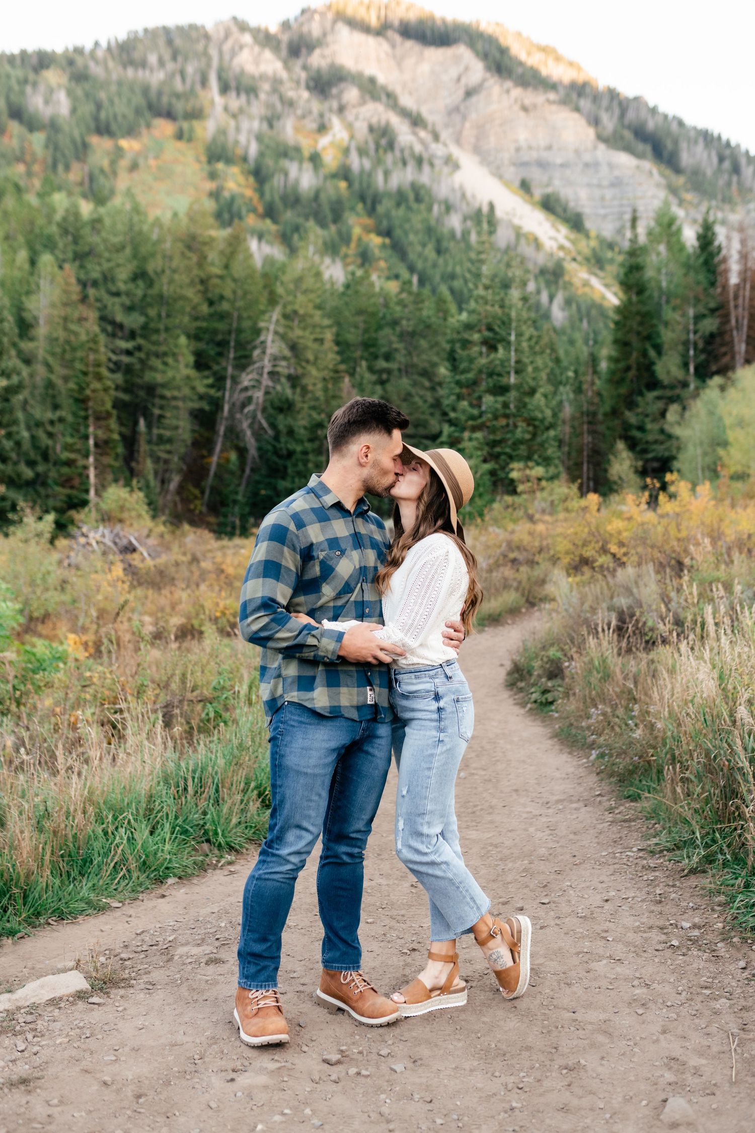 A couple shares a romantic moment on a hiking trail with mountains and evergreen trees in the background.
