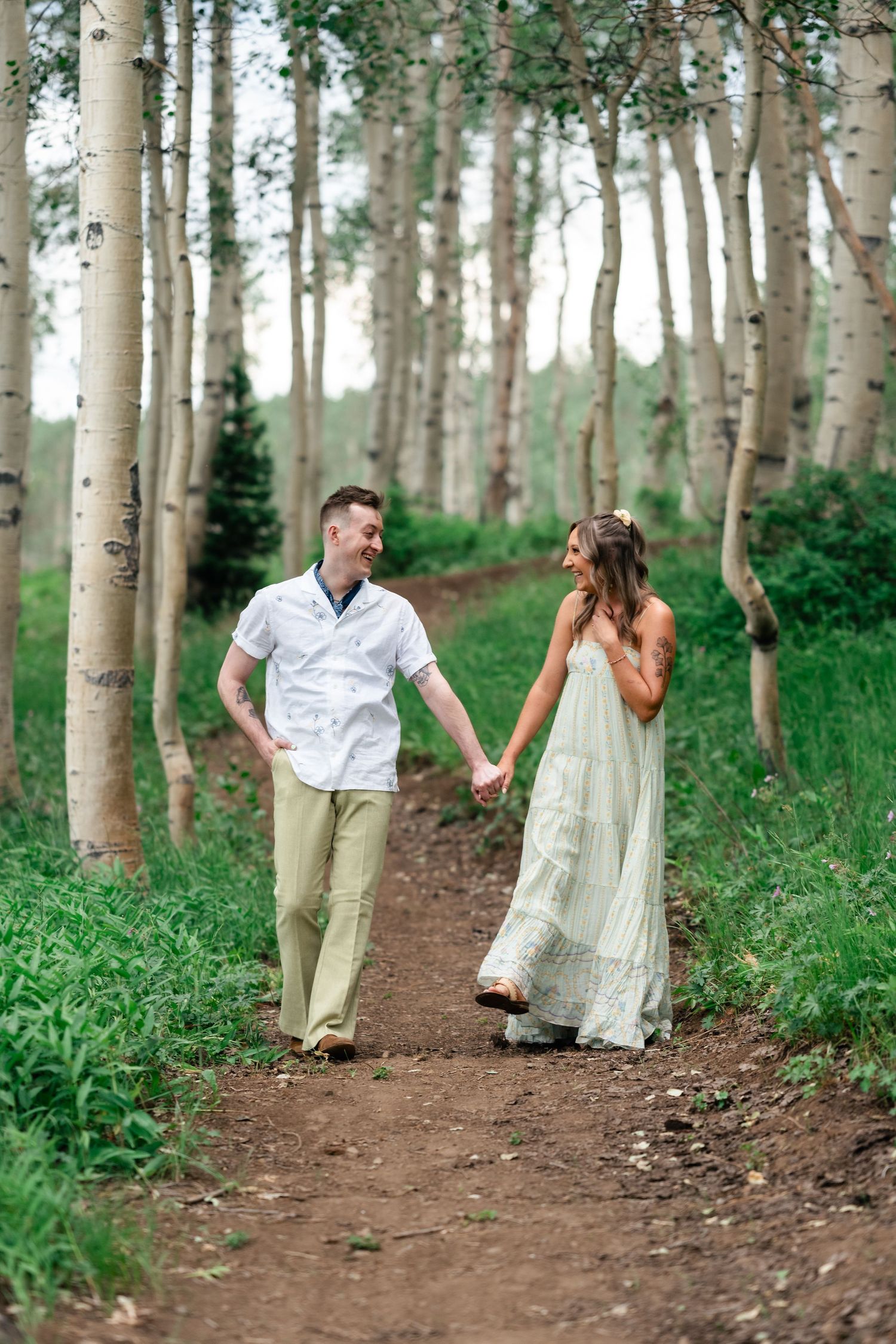 A couple walks hand in hand along a dirt path through a serene aspen forest on a summer day. Park City engagement photographer. Utah Wedding