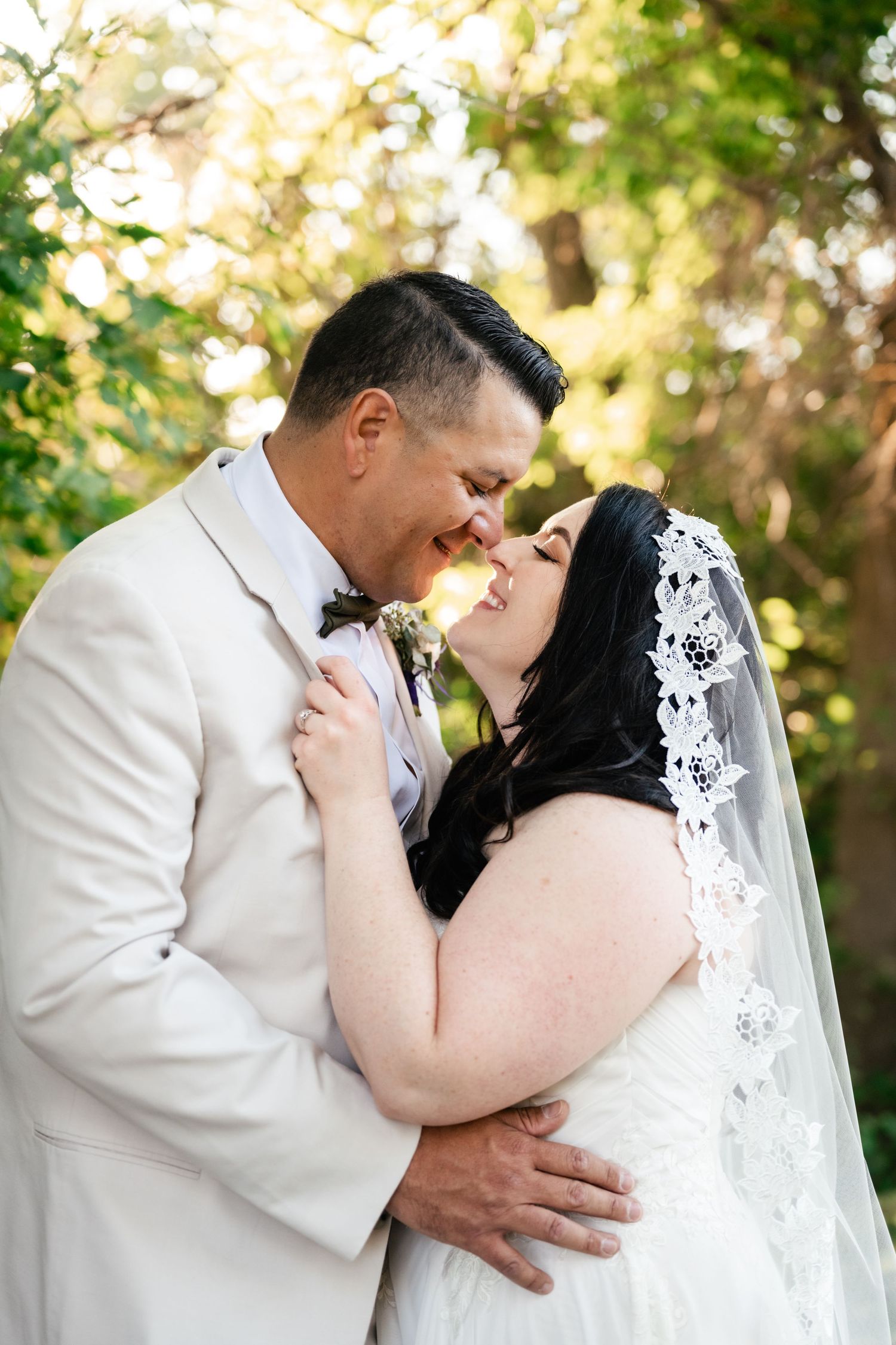 A romantic wedding moment captured outdoors with the bride in a lace veil and groom in cream suit embracing.