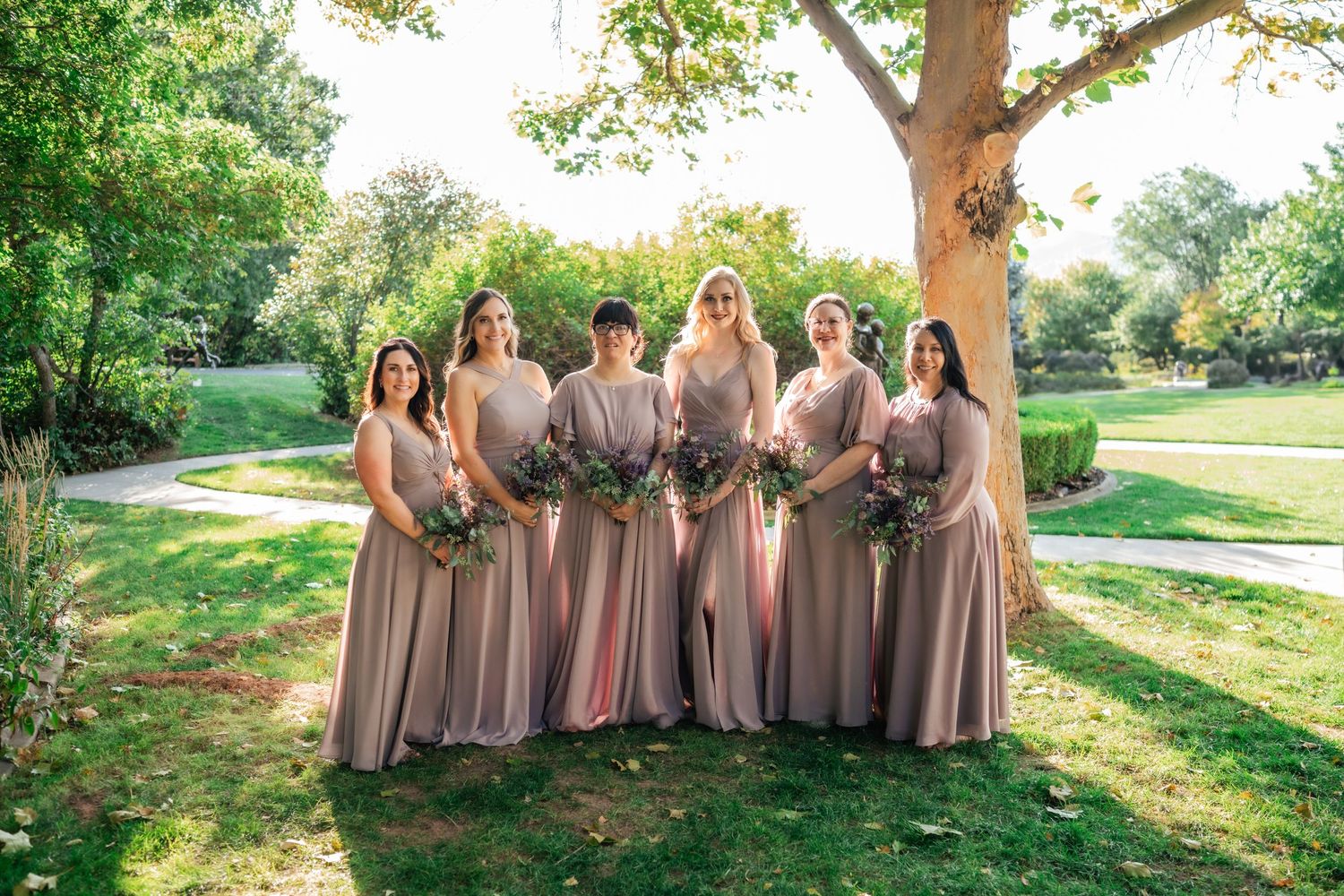 Wedding party members in dusty rose dresses stand together under a large tree in a sunlit garden setting.