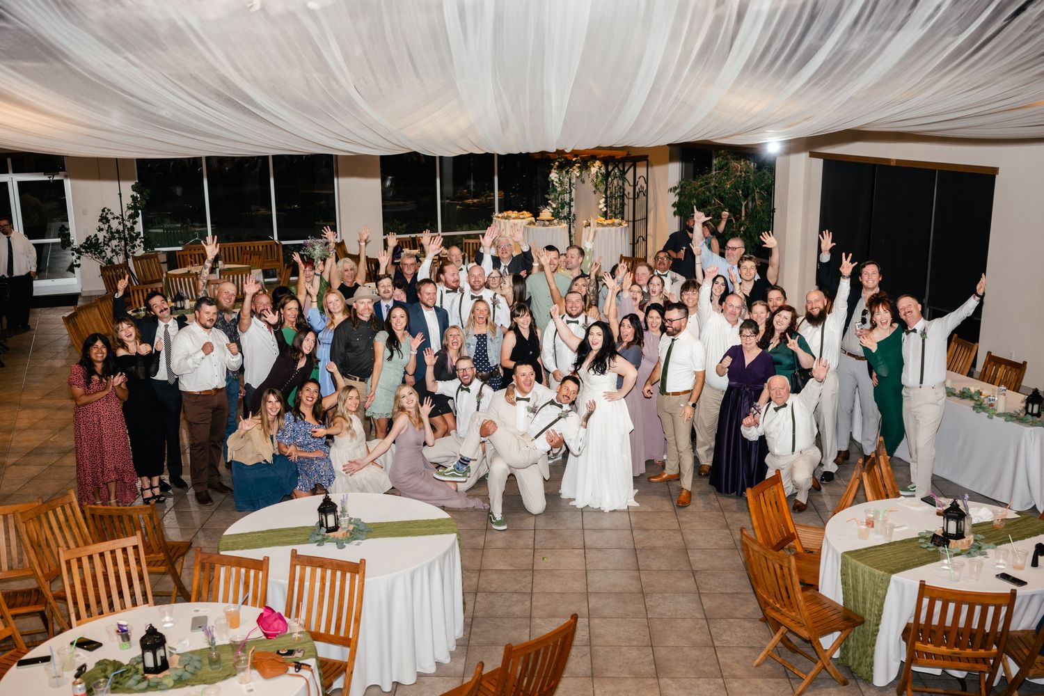 Large wedding reception group photo showing celebration with guests gathered around the newlyweds at an indoor venue.