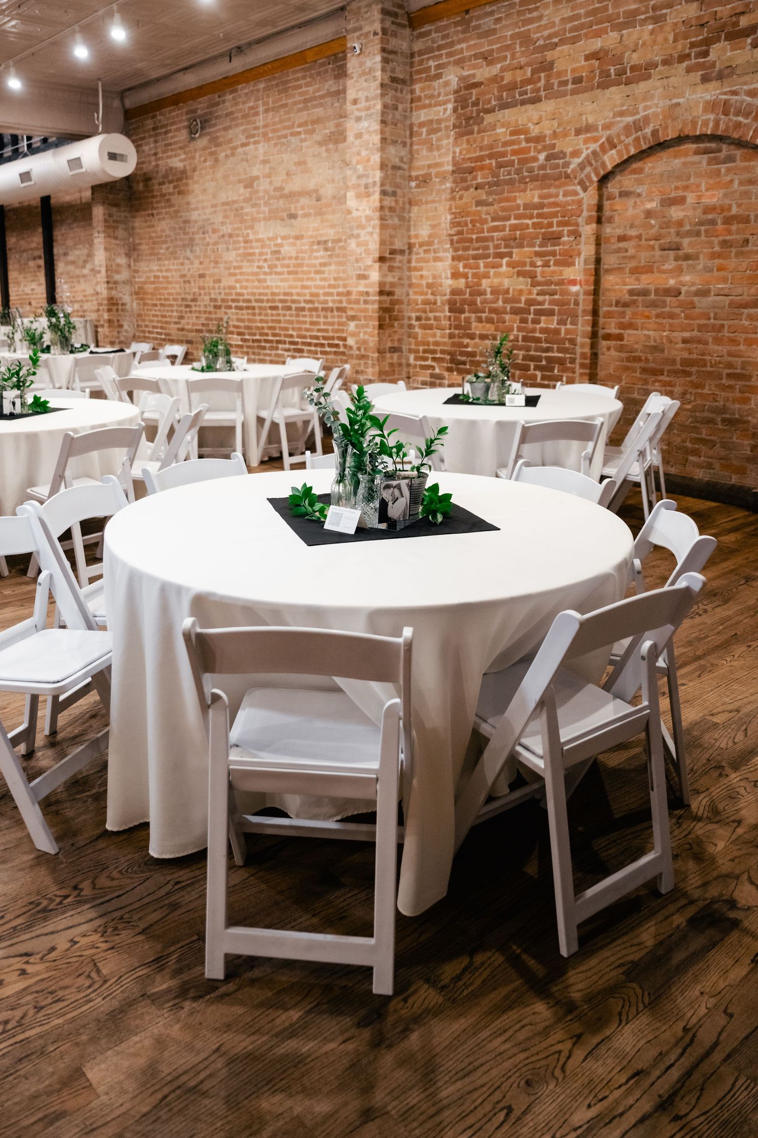 Round tables with white linens and greenery centerpieces arranged in an elegant venue with exposed brick walls.