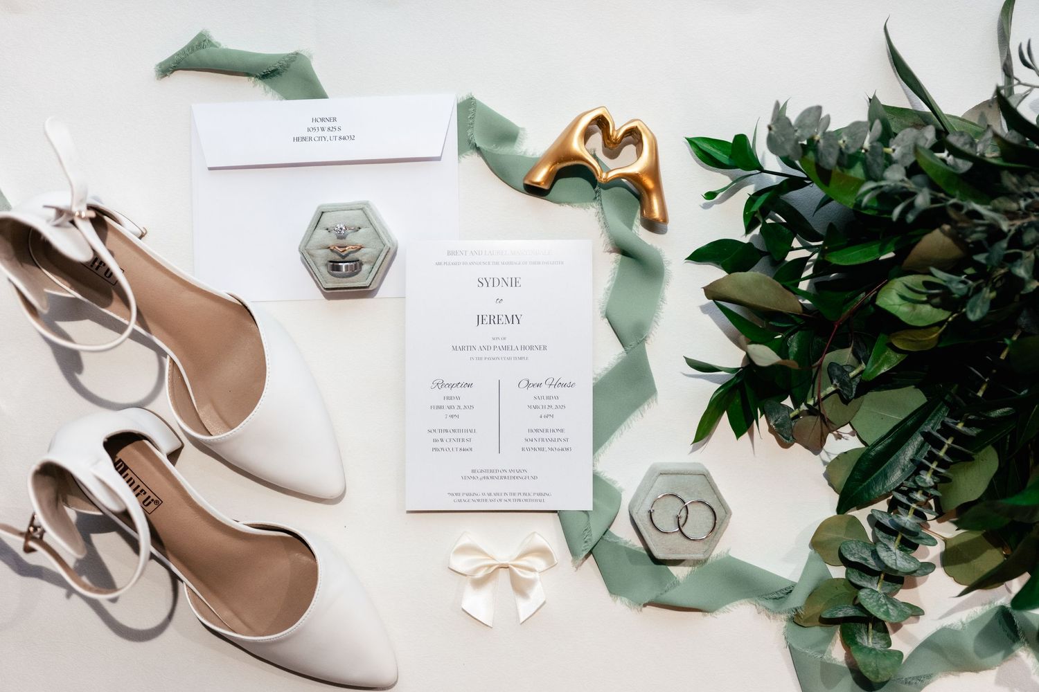 Wedding details flatlay with white shoes, invitation, rings, and green eucalyptus leaves on light background.