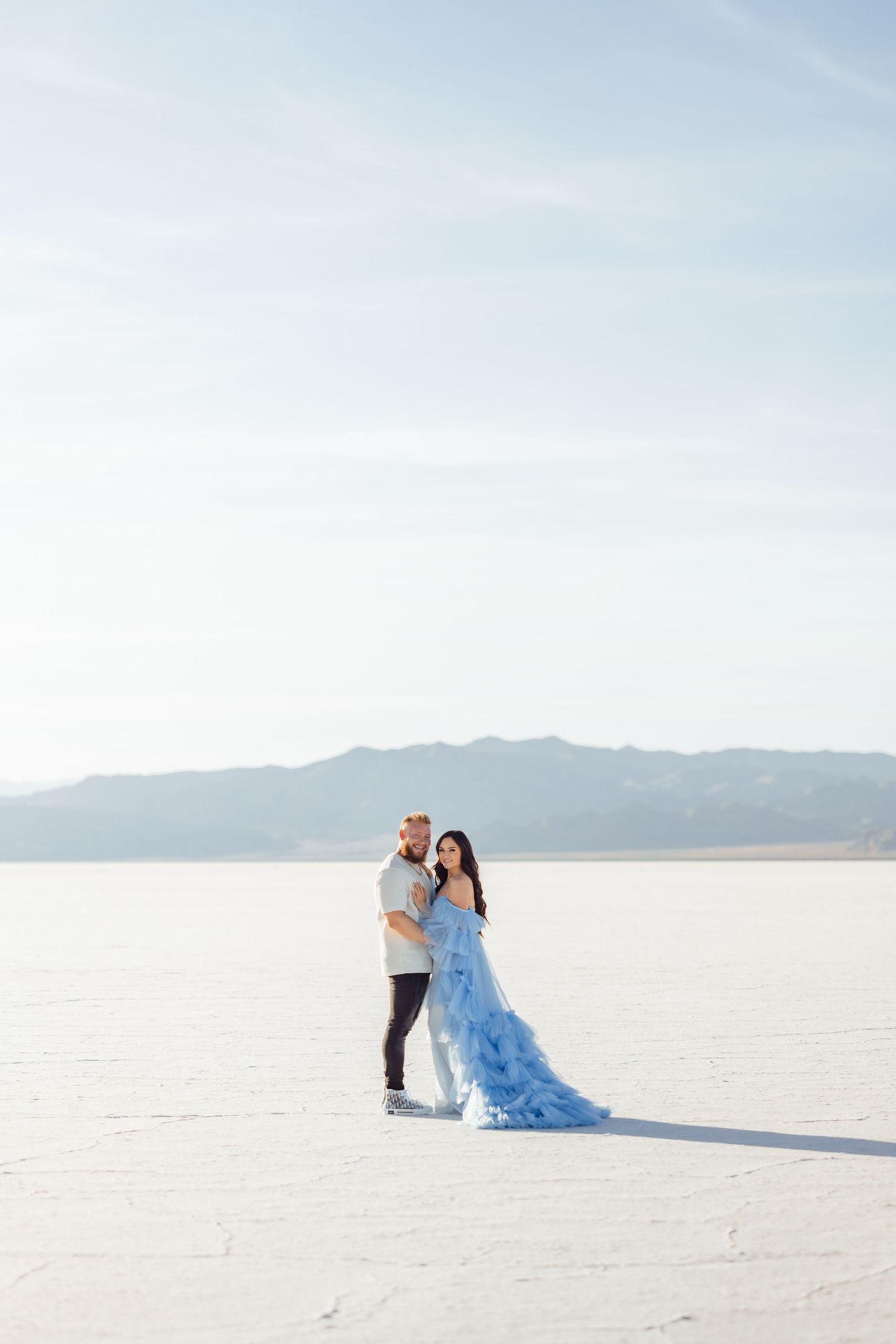 A couple embraces on a vast salt flat landscape with mountains in the background as the bride wears a flowing blue dress.