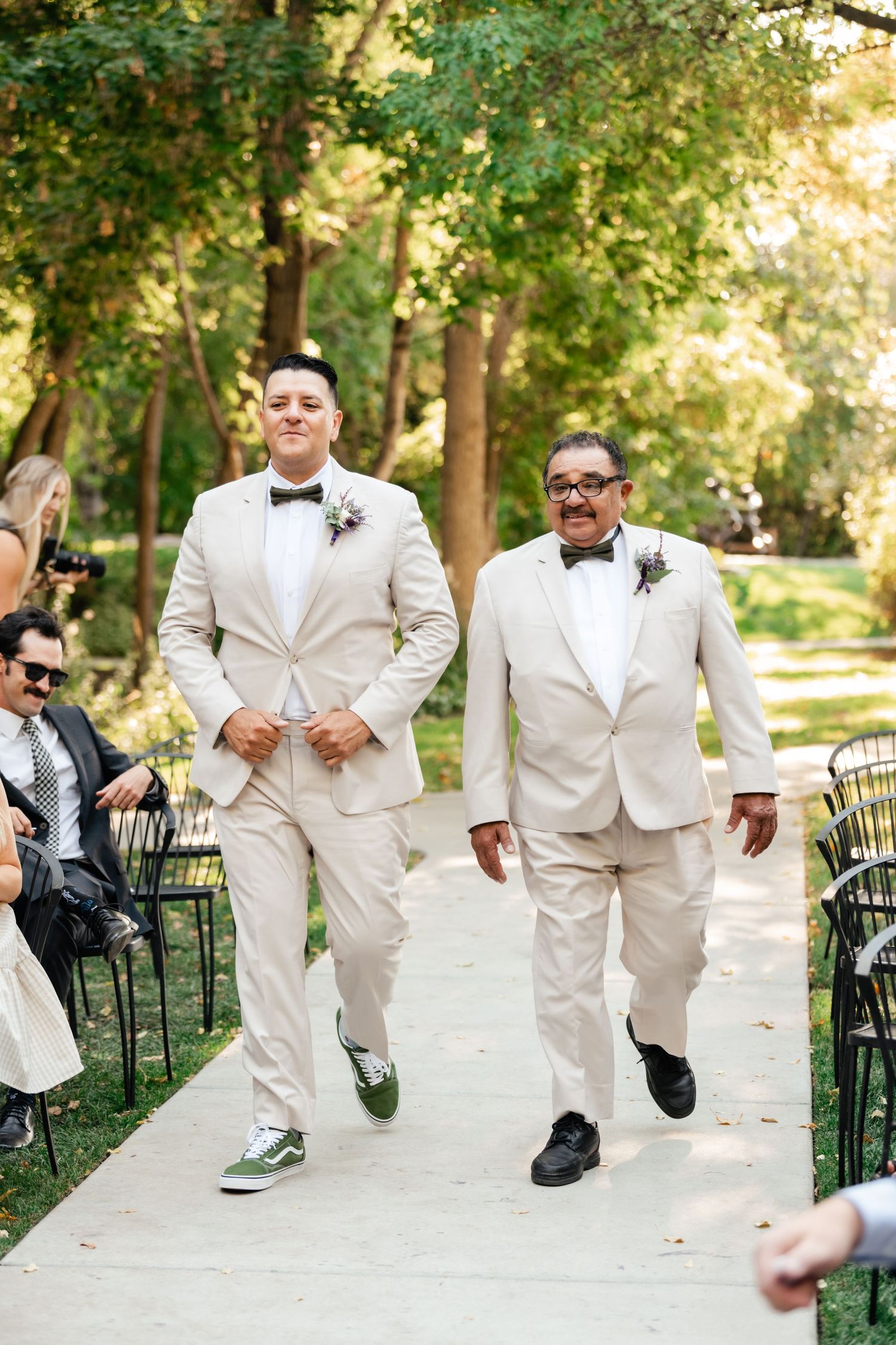 Two people wearing matching cream-colored suits and bowties walking down an outdoor garden path.
