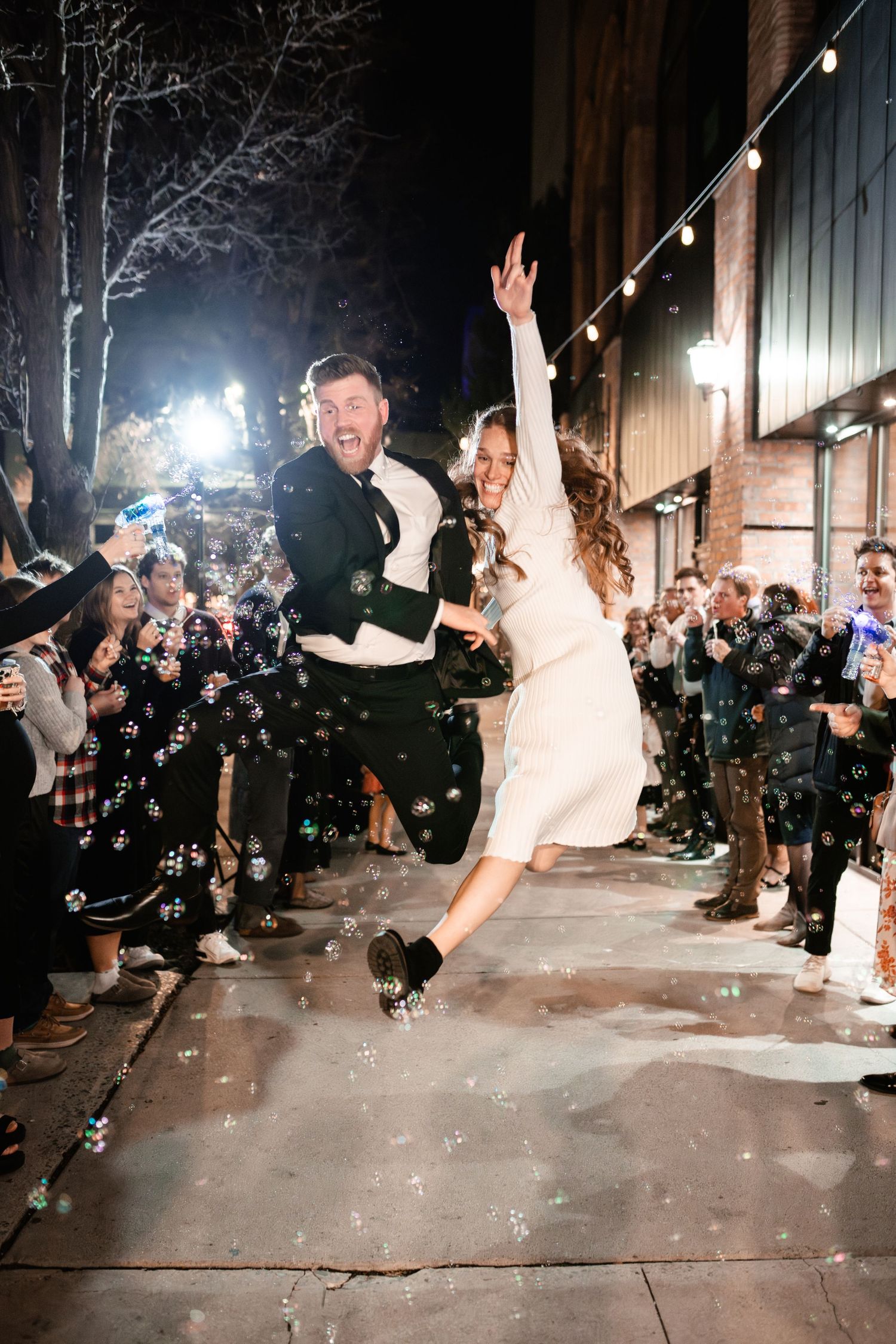 Joyful celebration as bubbles fall during a nighttime event with string lights and wooden barn backdrop.