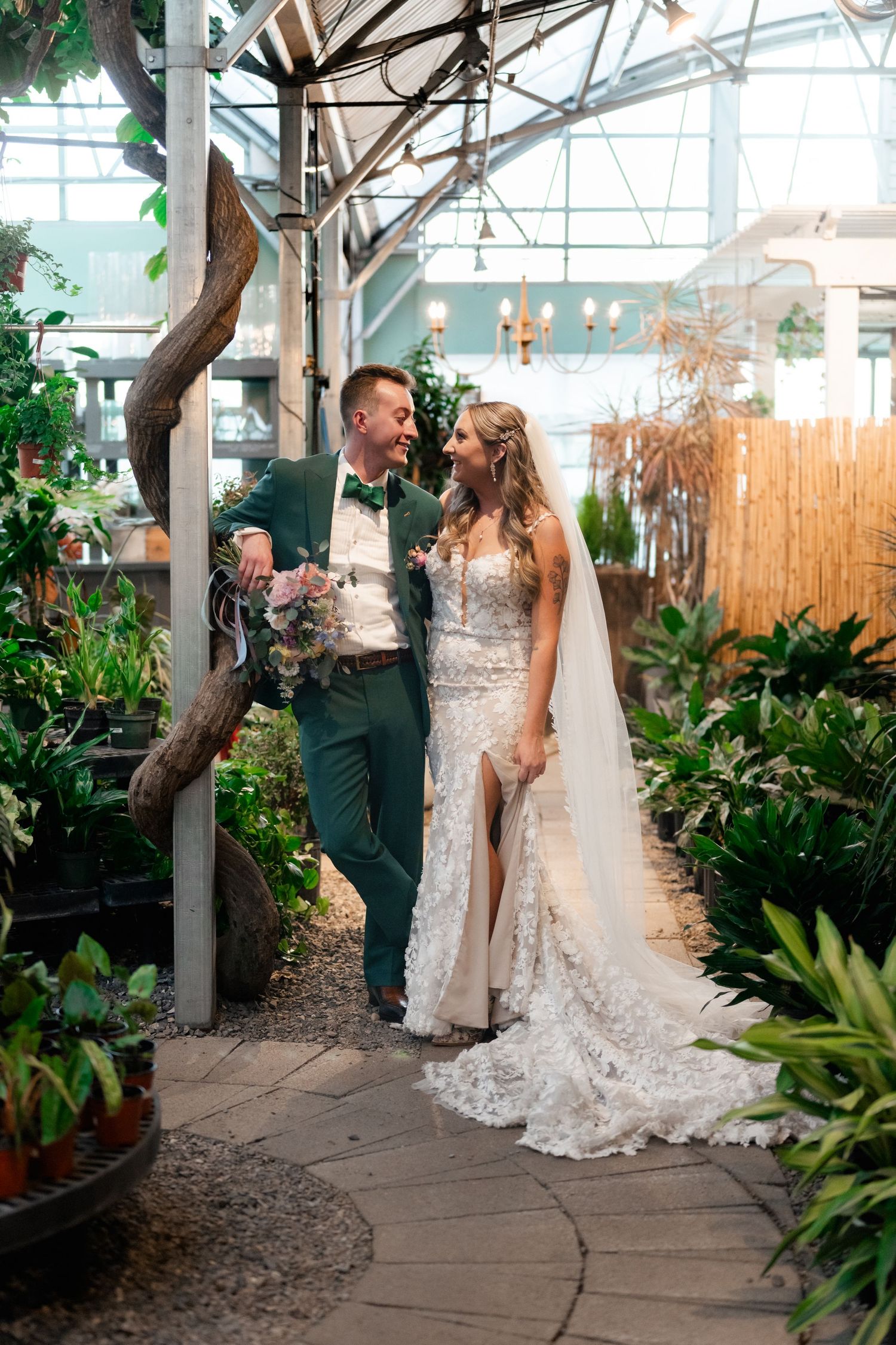 A romantic wedding couple stands together in a greenhouse conservatory surrounded by lush tropical plants and natural light.