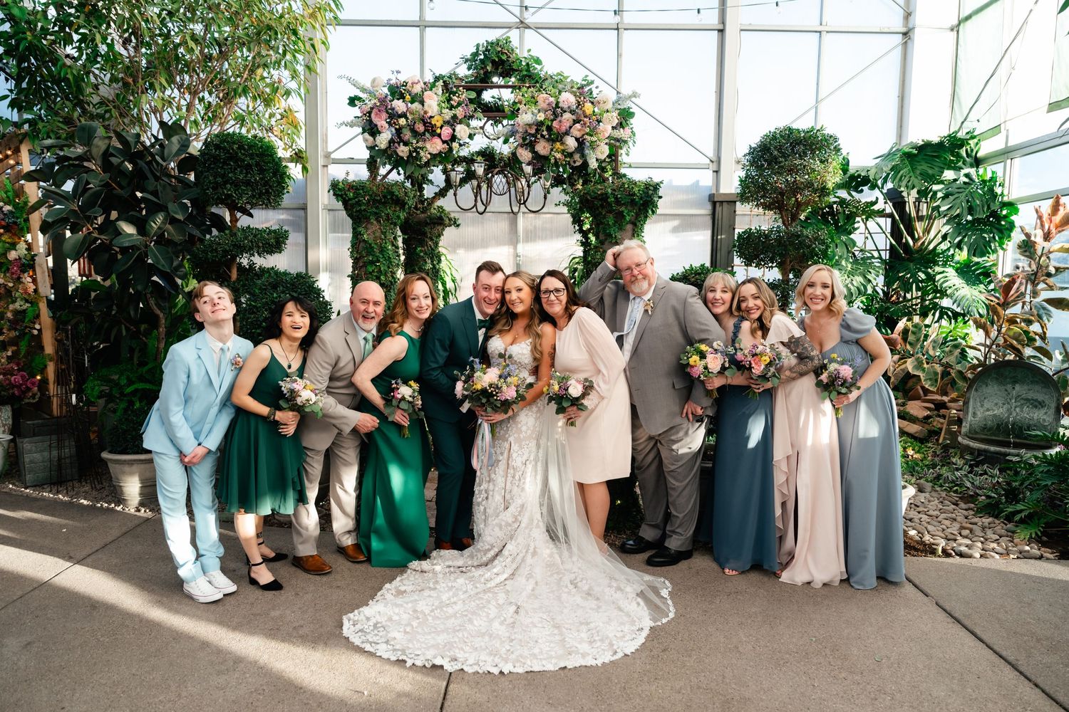 Wedding party poses together in a greenhouse setting with floral arch decorations and mixed emerald and neutral attire.