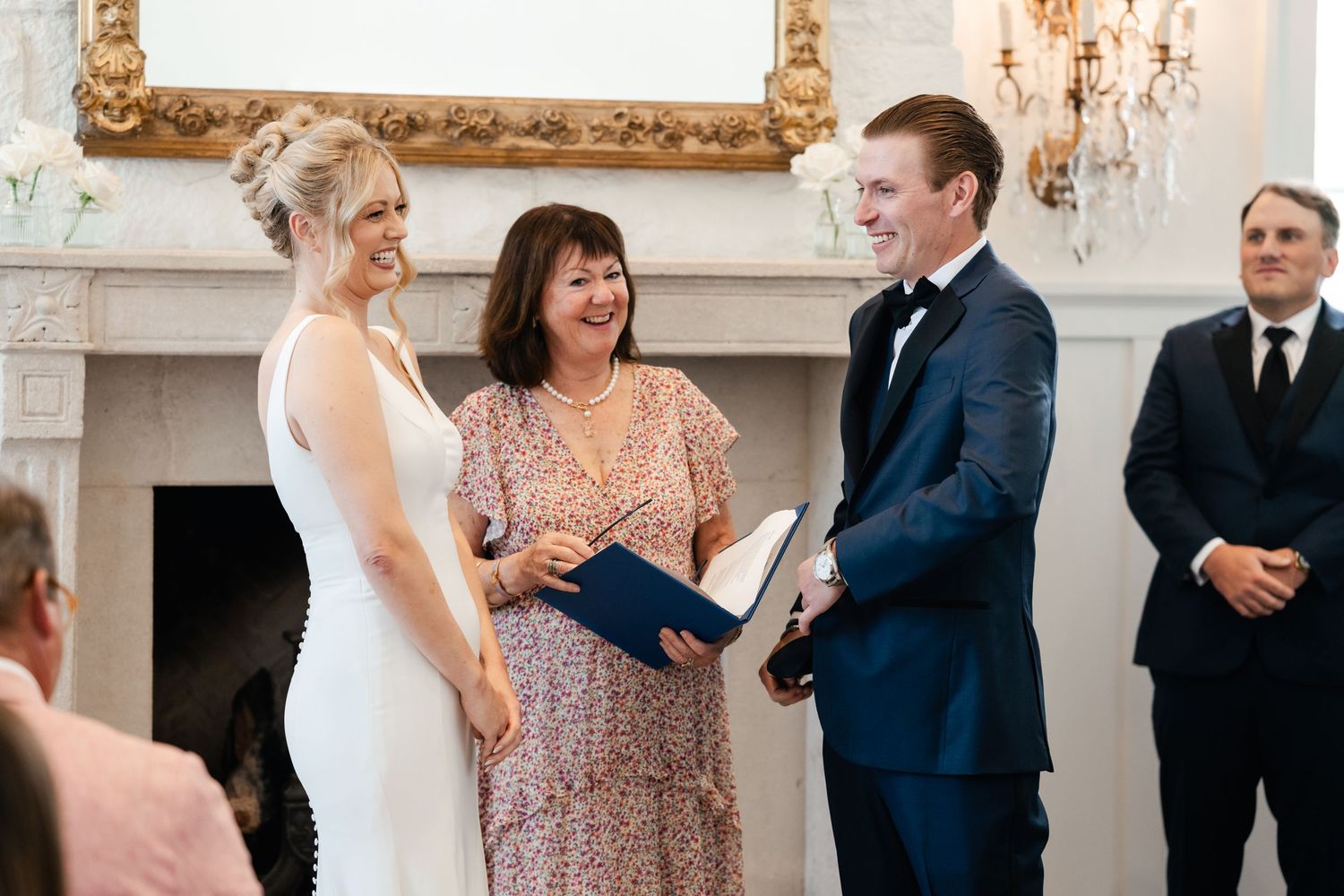 Wedding guests share joyful laughter during an elegant indoor ceremony with ornate gold-framed decor.