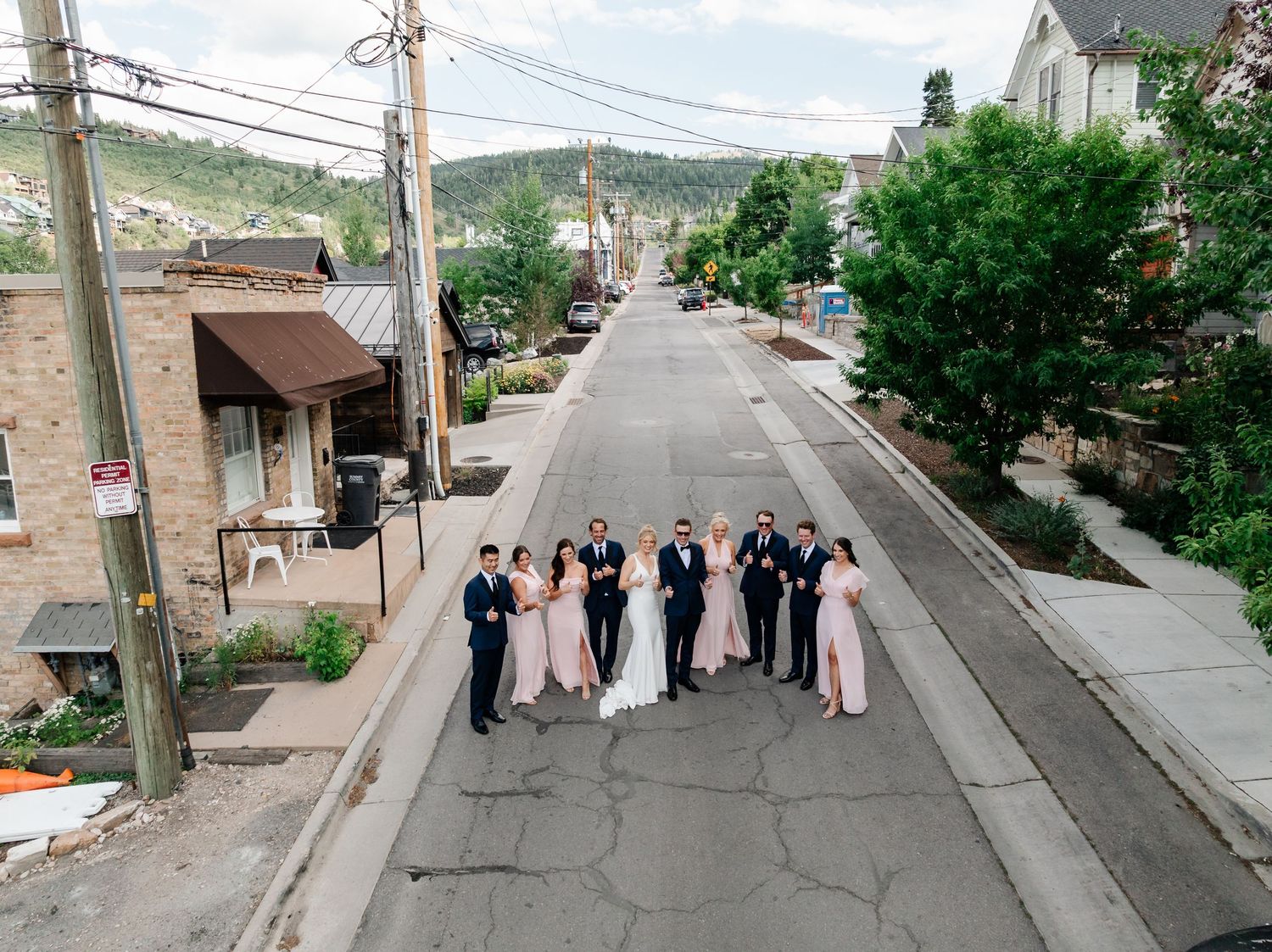 Wedding party in formal attire poses on a sloped residential street with trees and houses in the background.