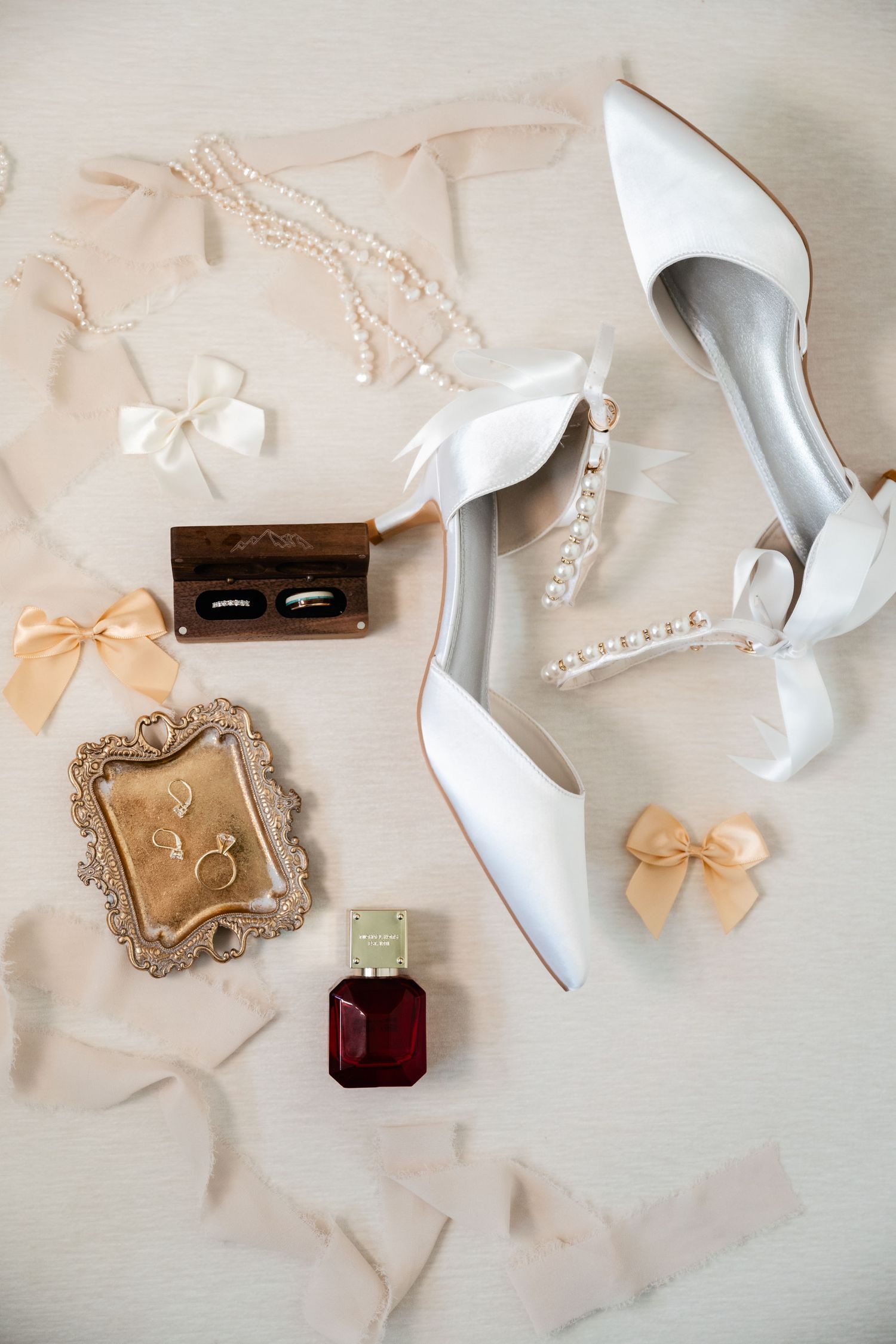 Wedding accessories including white heels, rings, perfume and delicate bows arranged on light fabric.