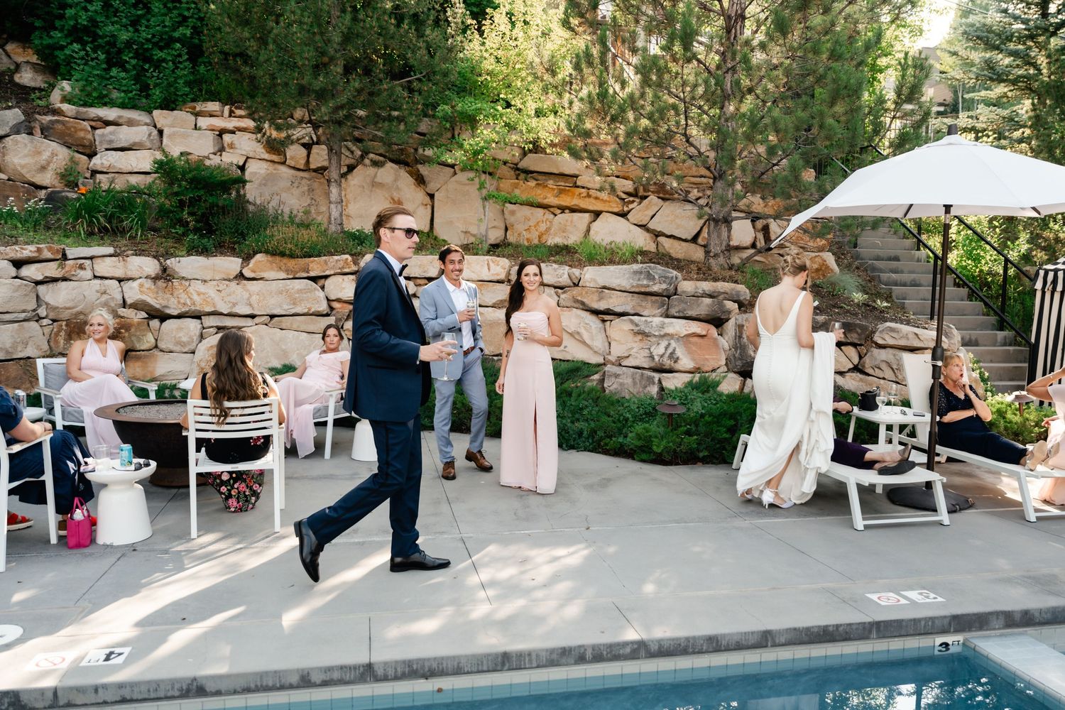 Wedding guests mingle on an outdoor patio with stone walls and white chairs.
