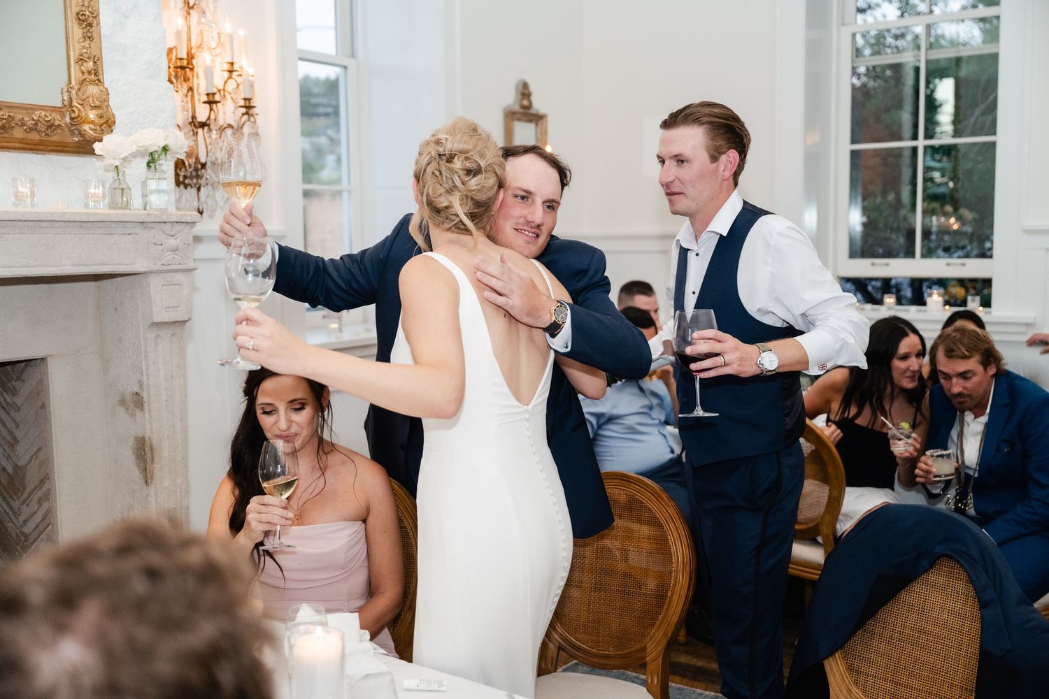 Elegant wedding reception with guests celebrating in bright white venue with chandeliers.