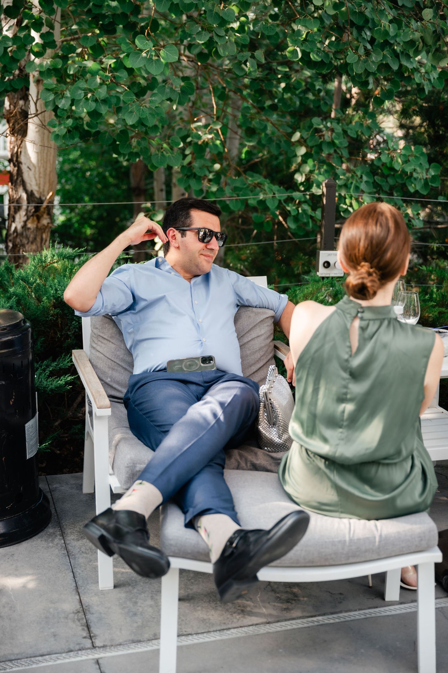 Two people relax together on a curved outdoor bench surrounded by lush green foliage on a sunny day.
