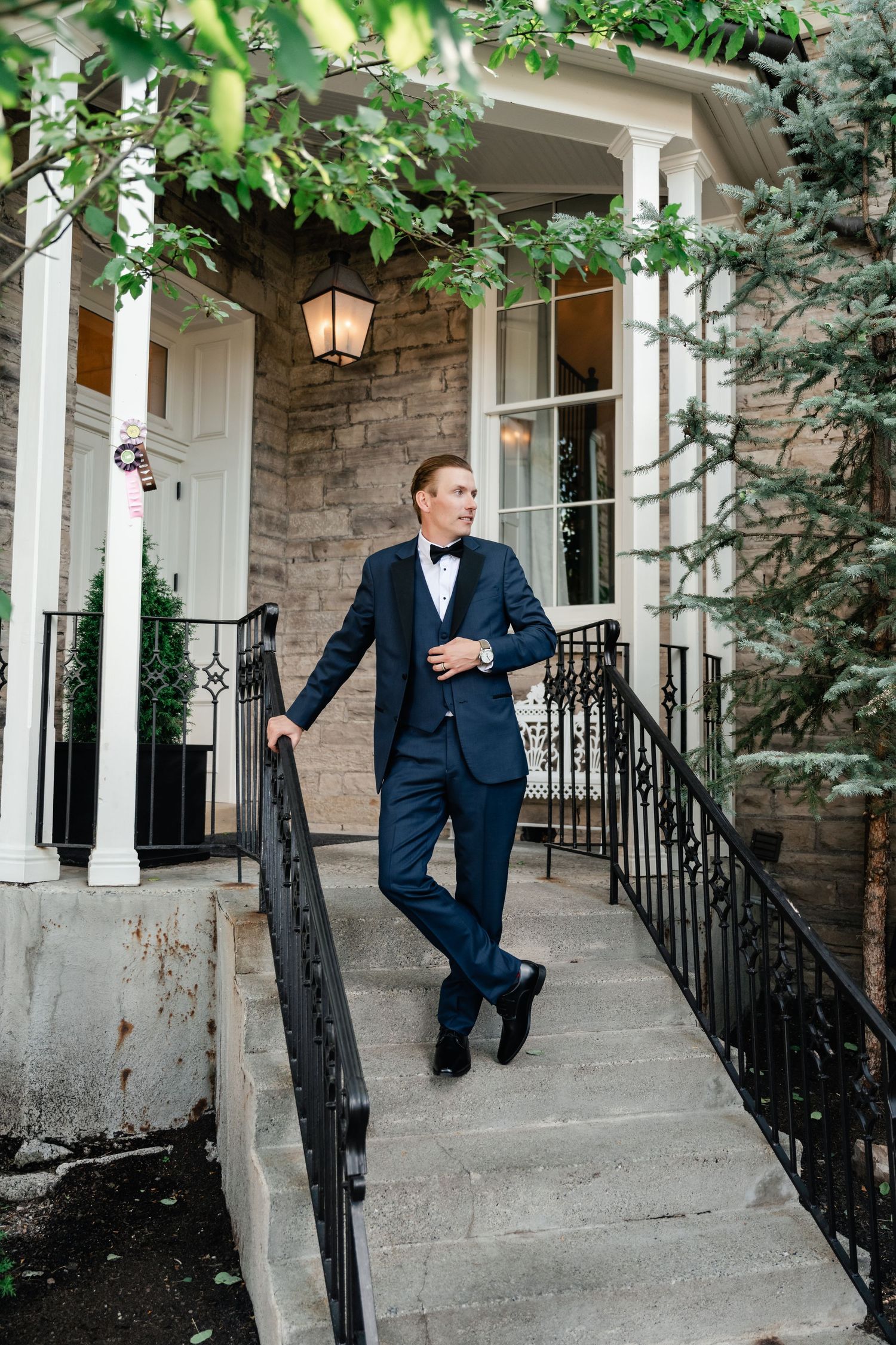A person in a navy suit poses playfully on stone steps with white columns and railings.