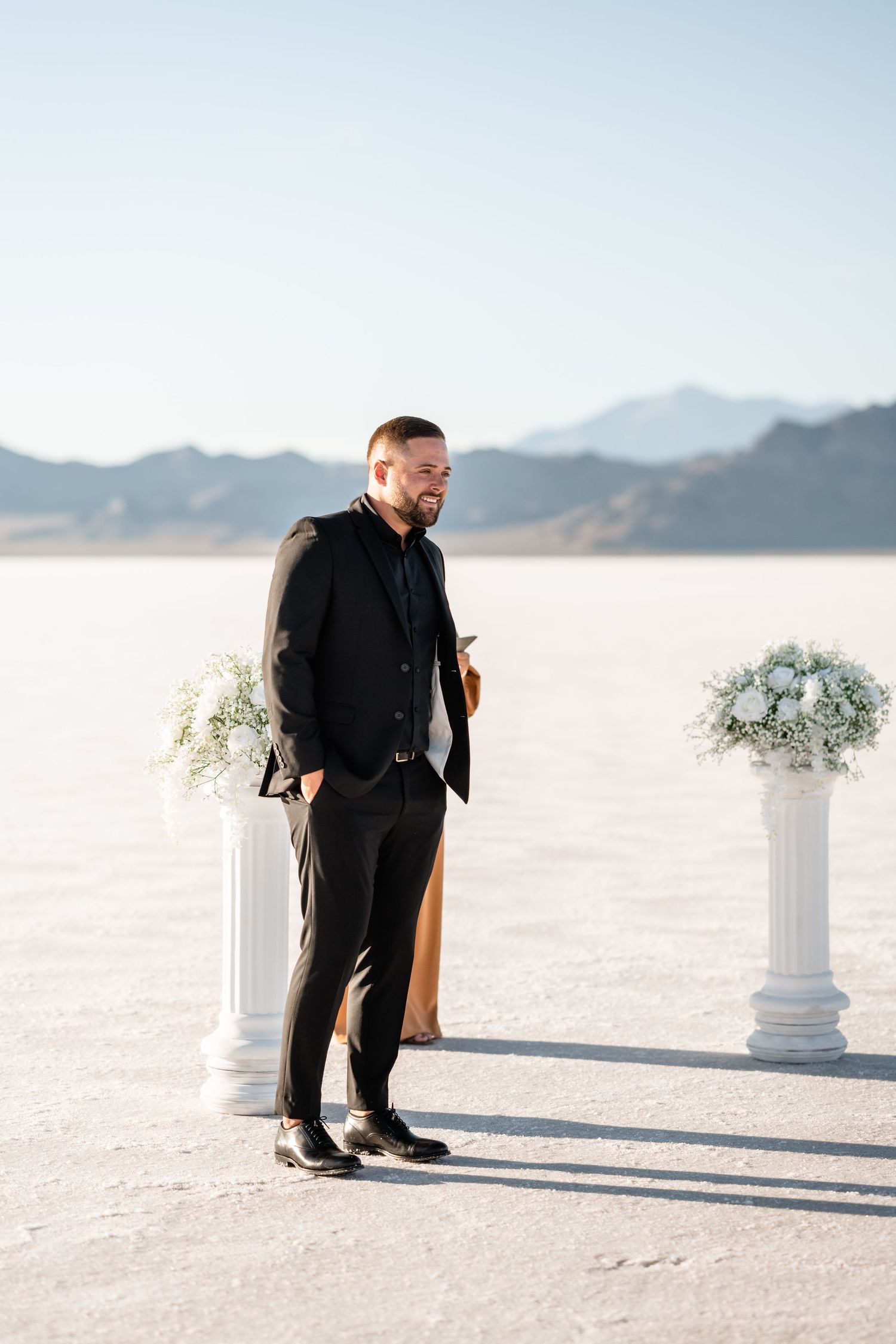 Groom in black suit waiting at outdoor ceremony site with white columns and mountain backdrop.