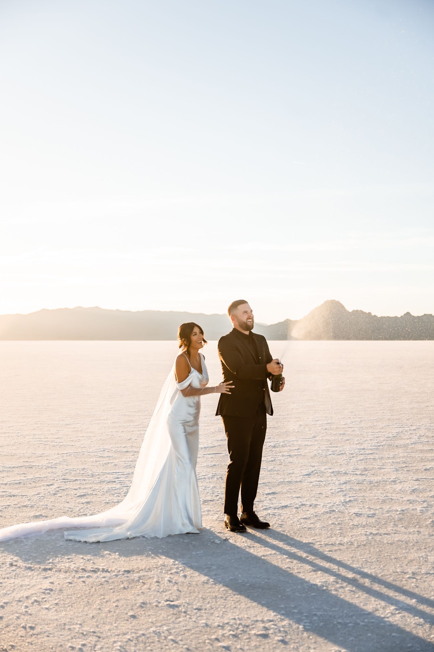A wedding couple poses together on white salt flats with mountains and sunset in the background.