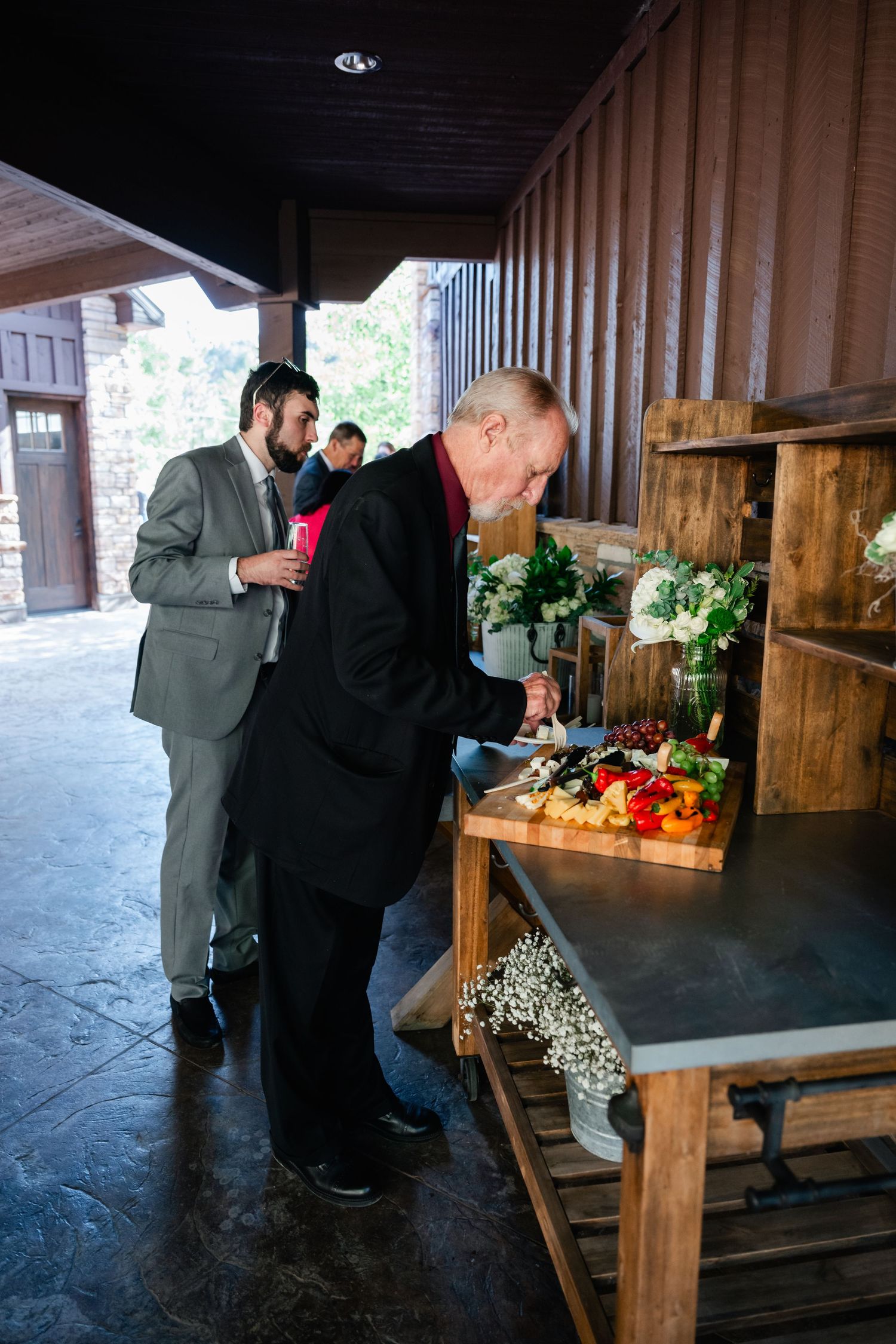People in formal attire examining floral arrangements on wooden tables at a rustic venue.