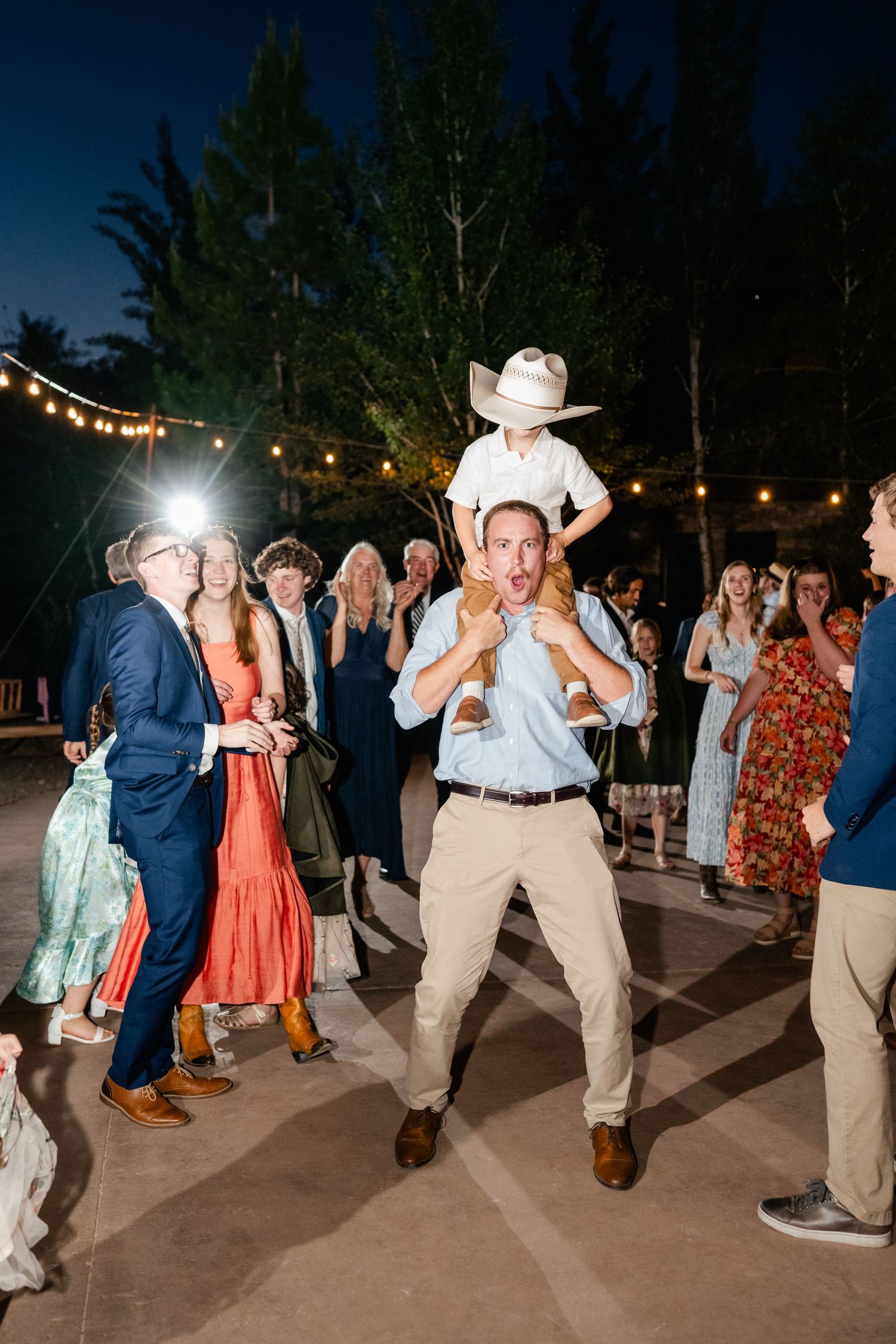 Wedding guests dance at an outdoor reception with string lights and trees in the background.