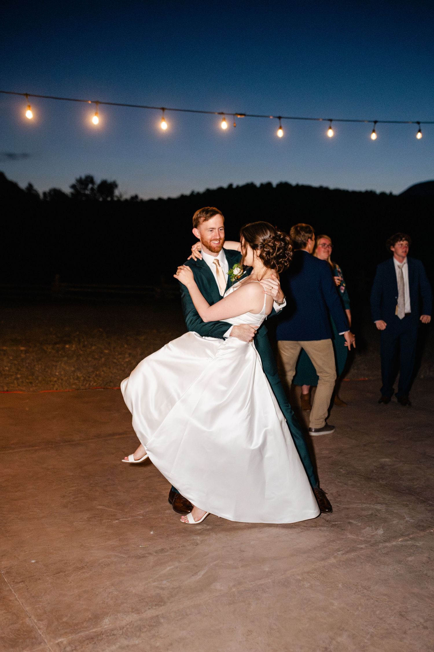 Wedding couple dances under string lights at dusk with guests watching in background.