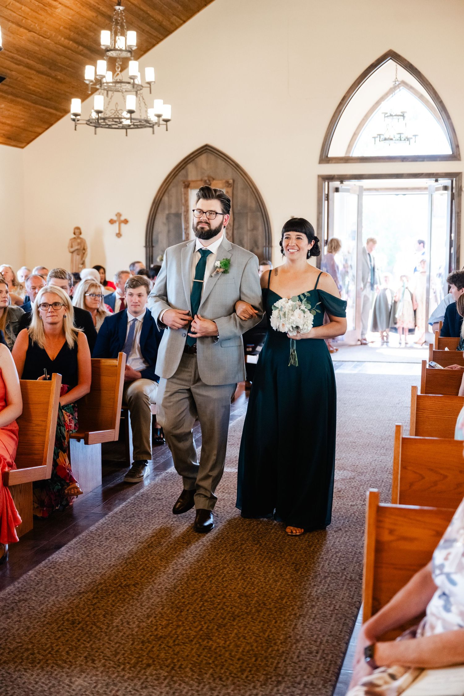 Wedding party members walk down the aisle in a church with gothic arched doorways and wooden ceiling.