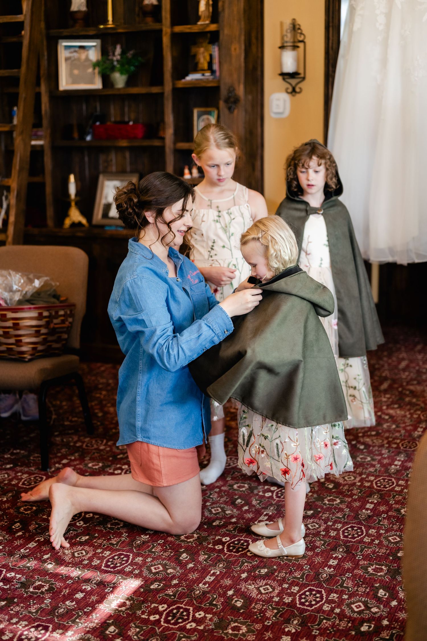 People in vintage-style clothing gather on a red patterned carpet in a cozy living room interior with wooden shelving.