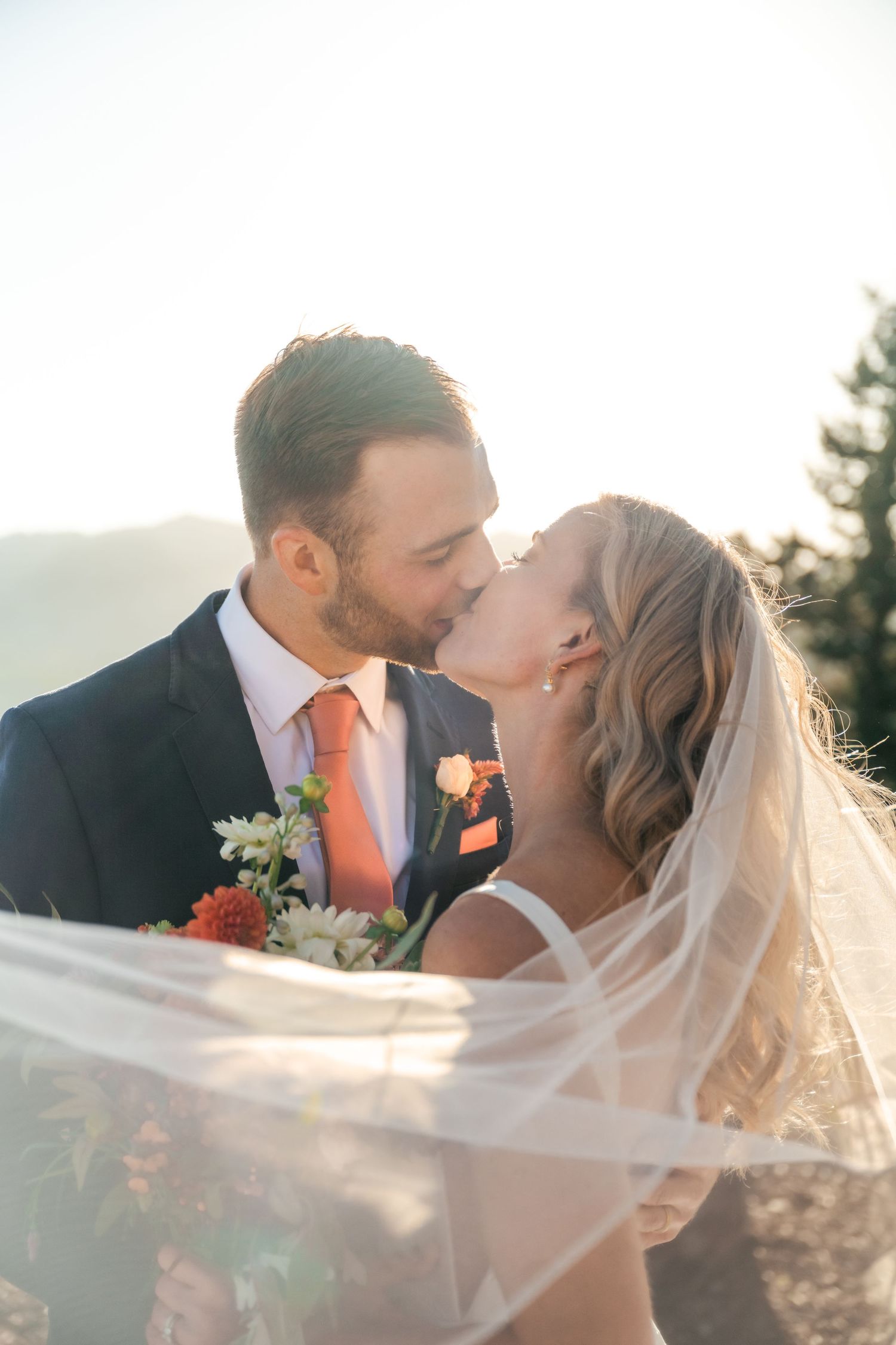 Intimate wedding kiss captured in golden mountain sunlight with flowing veil.