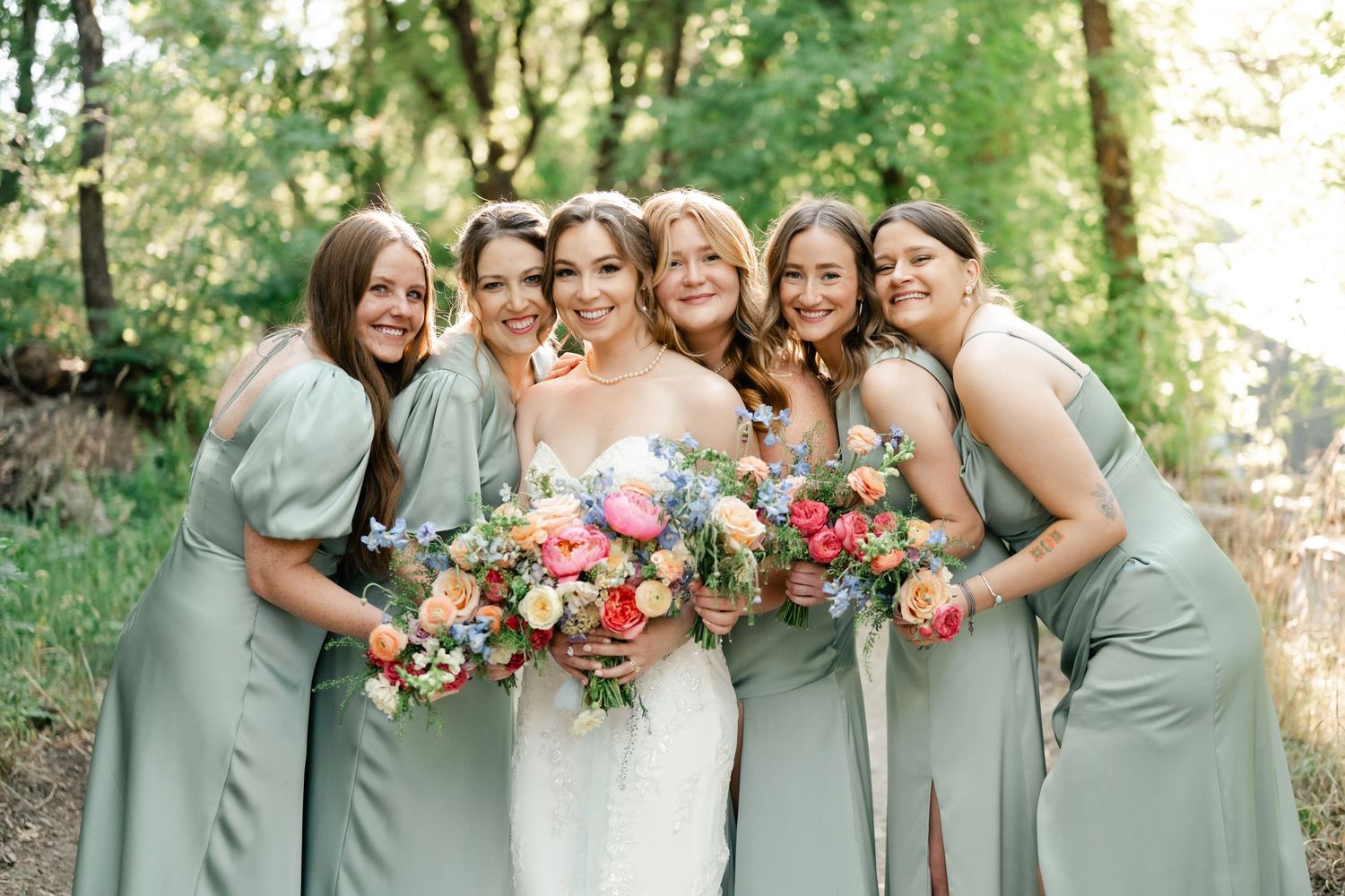 Bridesmaids in sage green dresses pose together holding pink and red bouquets in a sunny outdoor setting. Park City Wedding Photographer.