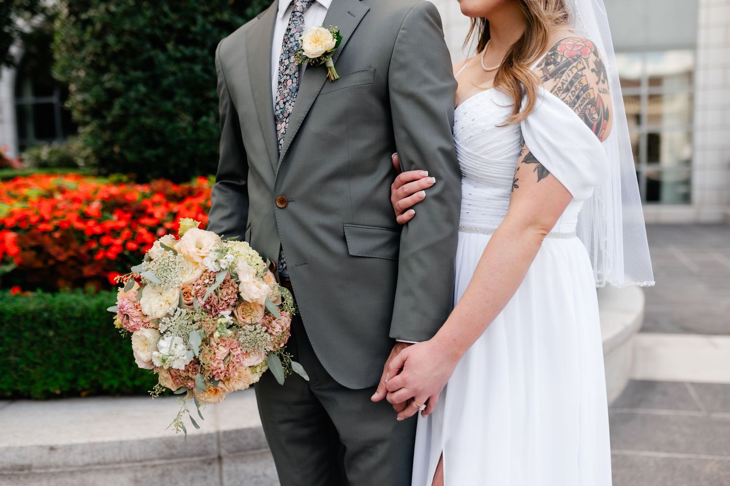 A bride in white dress and groom in gray suit stand together with a blush pink and cream floral bouquet.