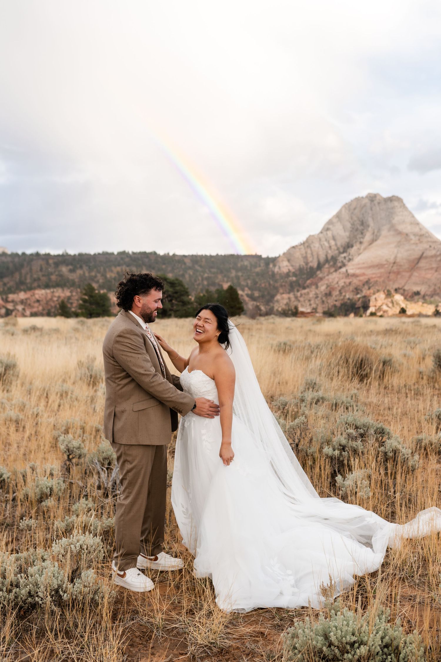 A romantic wedding photoshoot in a scenic mountain field with flowing white dress and brown suit at sunset.