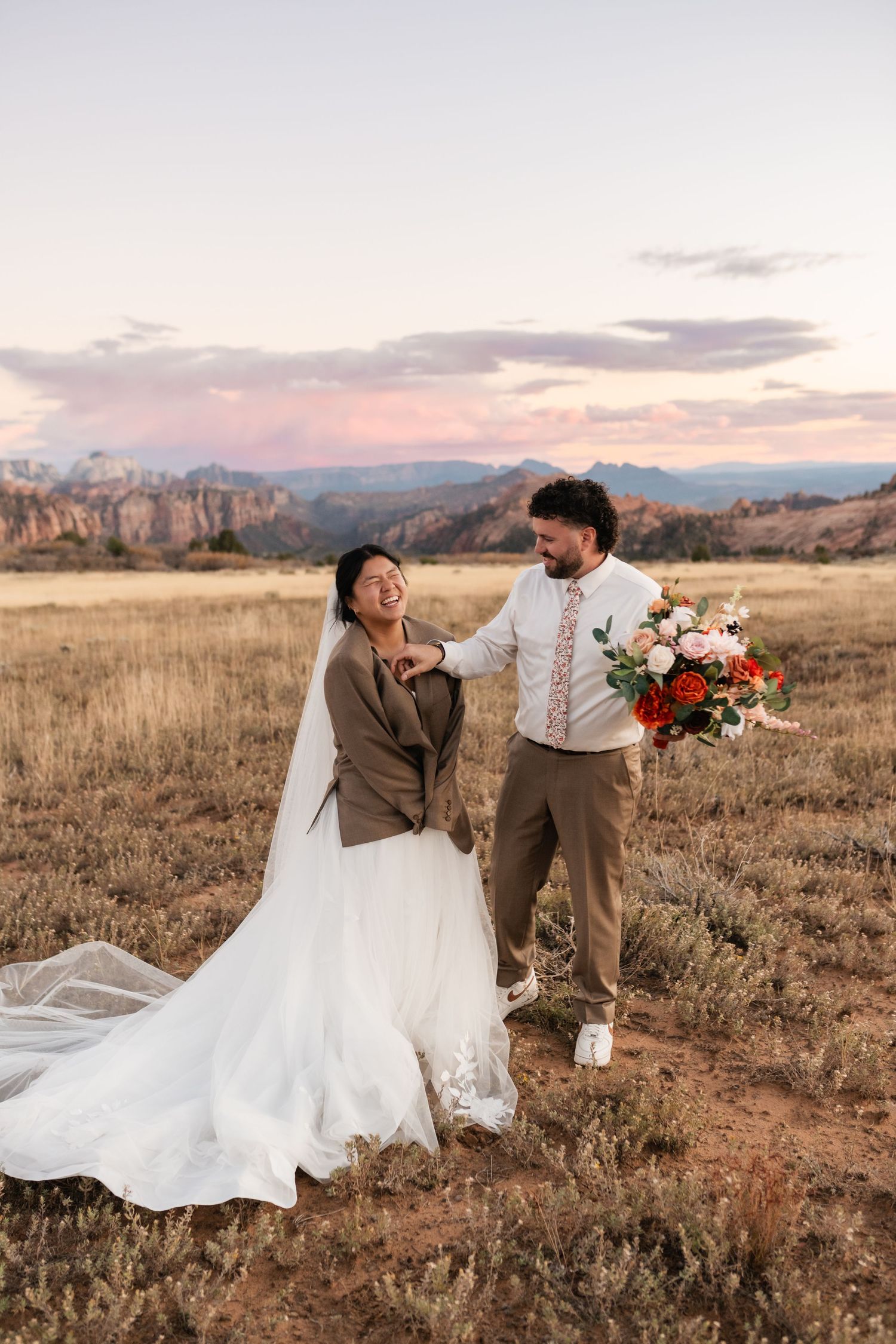 Wedding couple shares joyful moment in desert field with mountain backdrop at dusk.