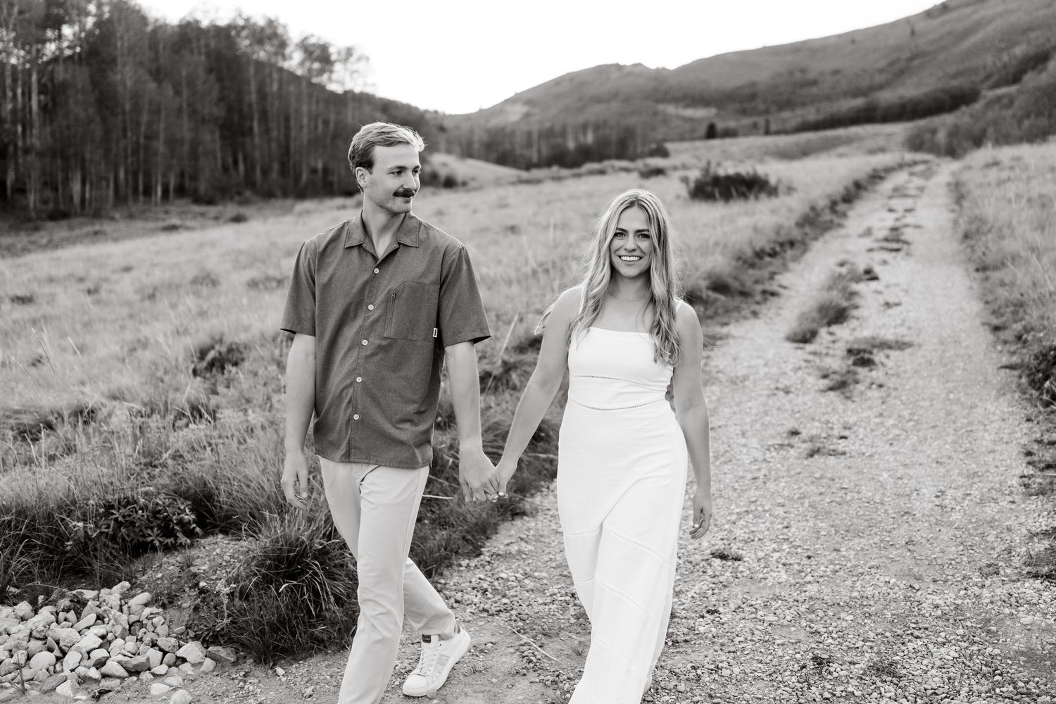 Couple holding hands walking down a dirt path in a mountain meadow, black and white engagement photo.
