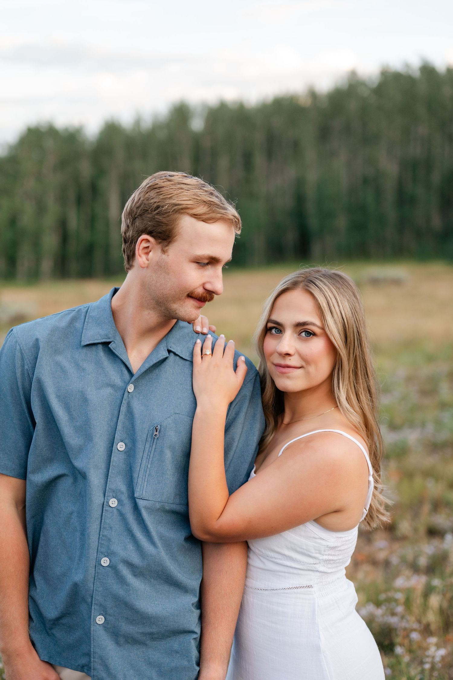 Couple standing in a meadow, woman resting hand on mans shoulder as he looks down at her tenderly after Park City surprise proposal.