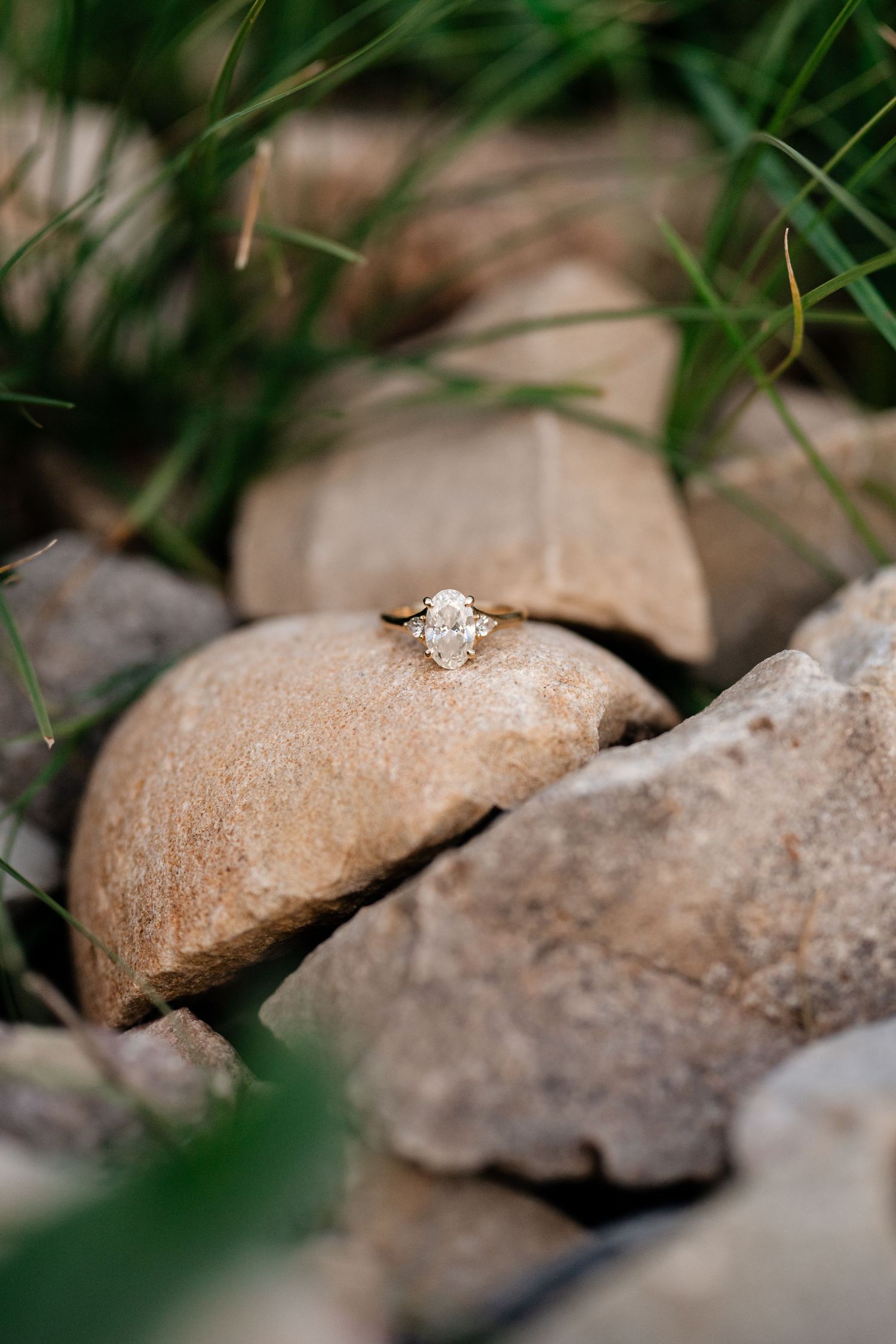 Gold engagement ring with oval diamond solitaire resting on natural rocks surrounded by green grass after Park City surprise proposal.