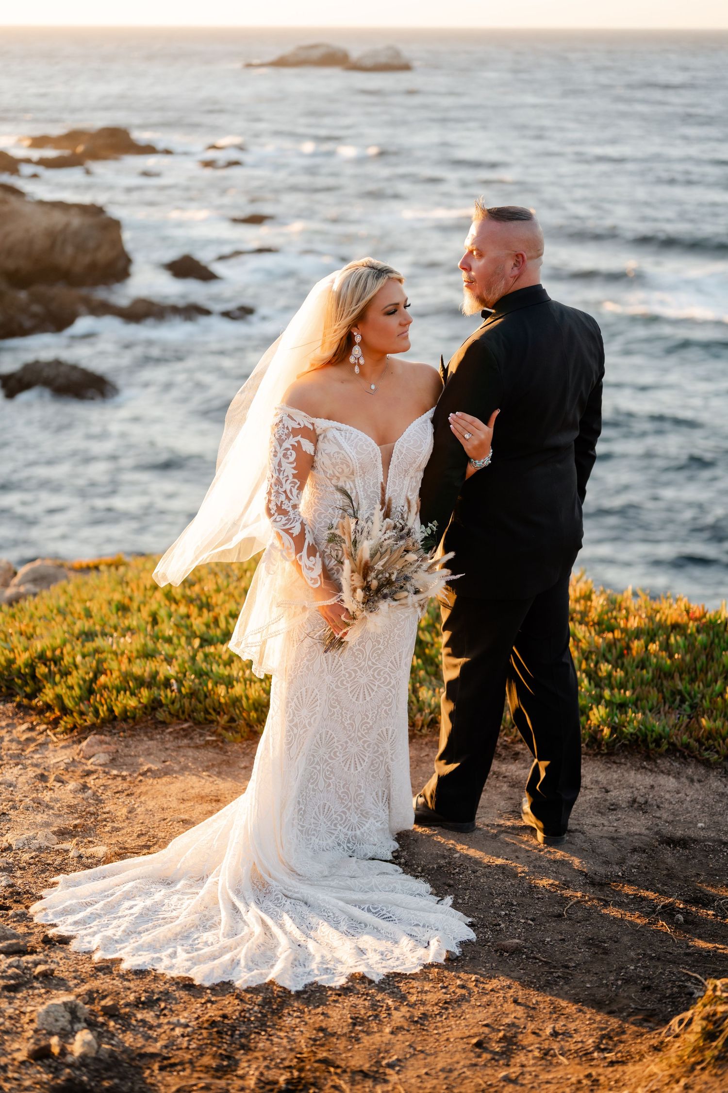 A coastal wedding portrait at sunset with dramatic rocky cliffs and ocean waves in the background.