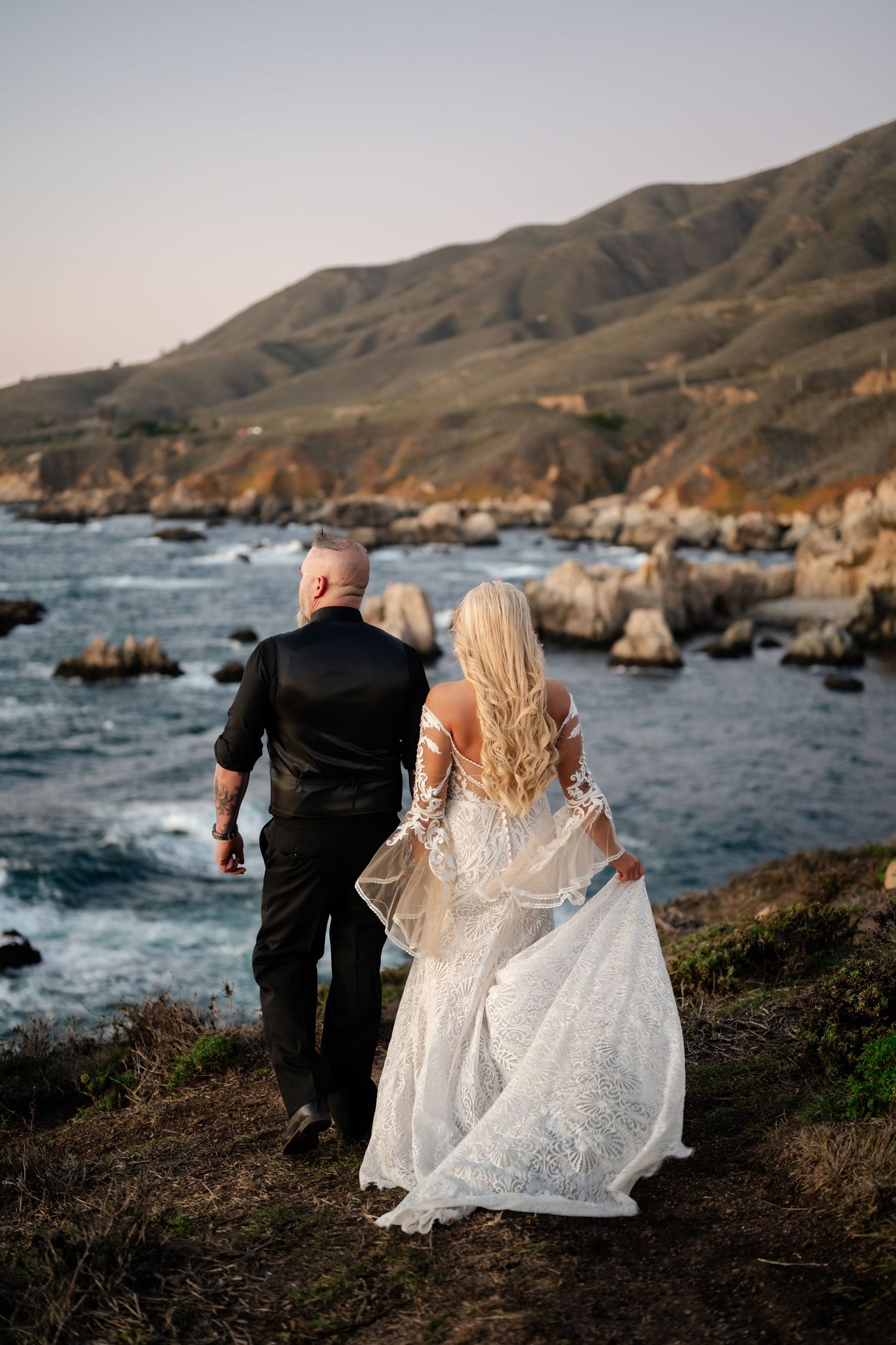 A couple walking along the rugged Big Sur coastline with crashing waves and rocky shores in the background.
