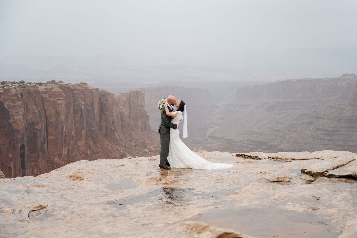 Dramatic wedding photography session on edge of steep canyon cliffs with expansive desert vista.
