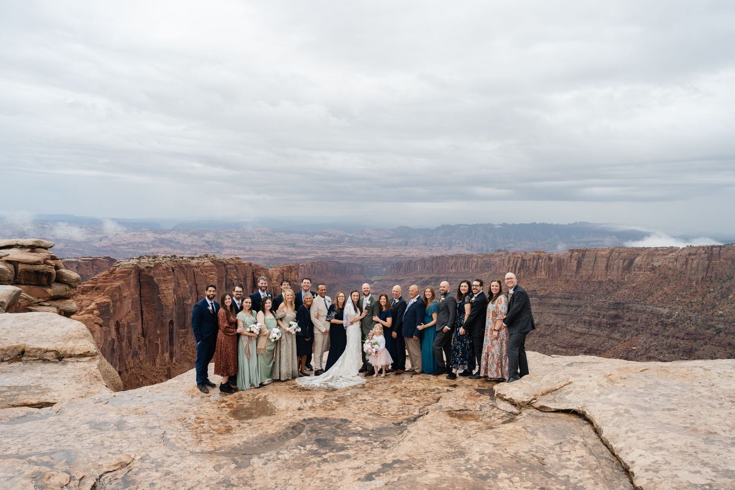 Large wedding party gathered on scenic desert overlook with dramatic canyon landscape stretching into distance.