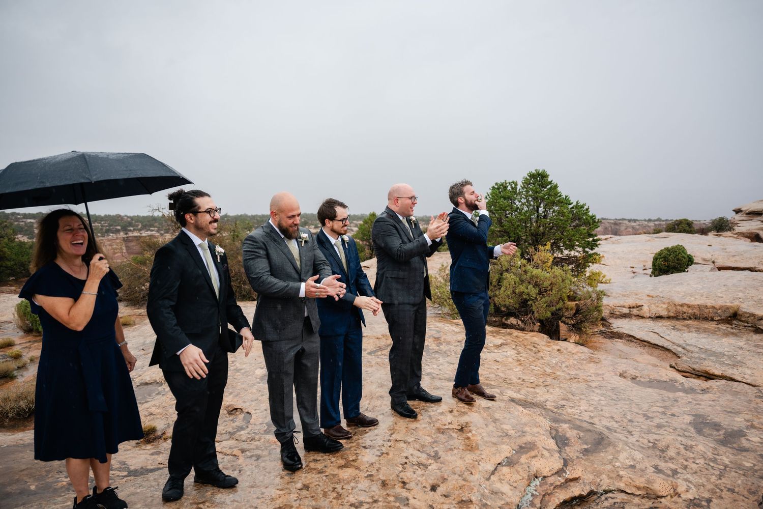 Wedding guests with umbrellas observe ceremony on rocky desert overlook.